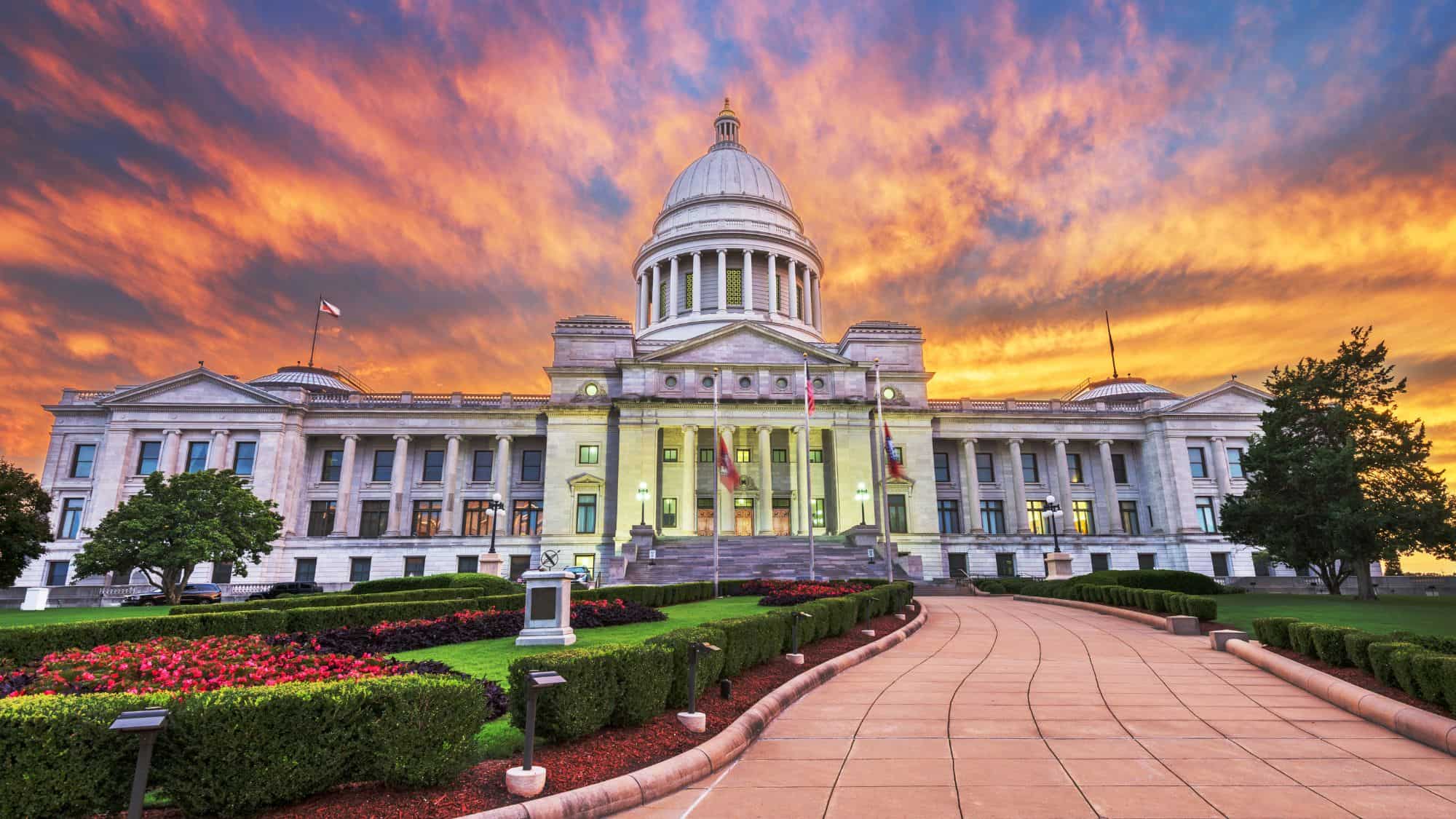 The Arkansas State Capitol building glows under a dramatic orange and purple sky, with a manicured path and gardens leading to its grand entrance.