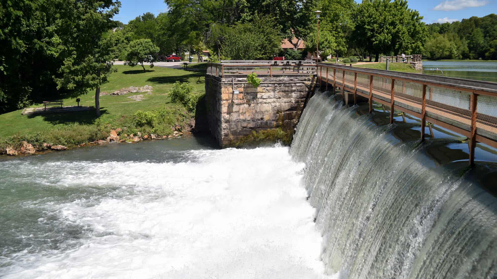 A small dam spills gently over a stone wall into a foamy stream, bordered by green lawns, trees, and a park walkway above.