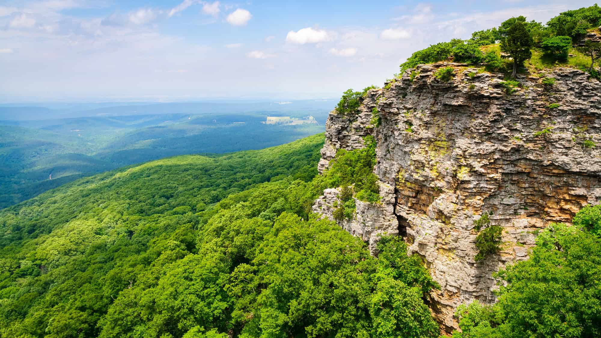 A rugged bluff juts out from dense forest, offering sweeping views across layers of rolling green hills beneath a bright blue sky.