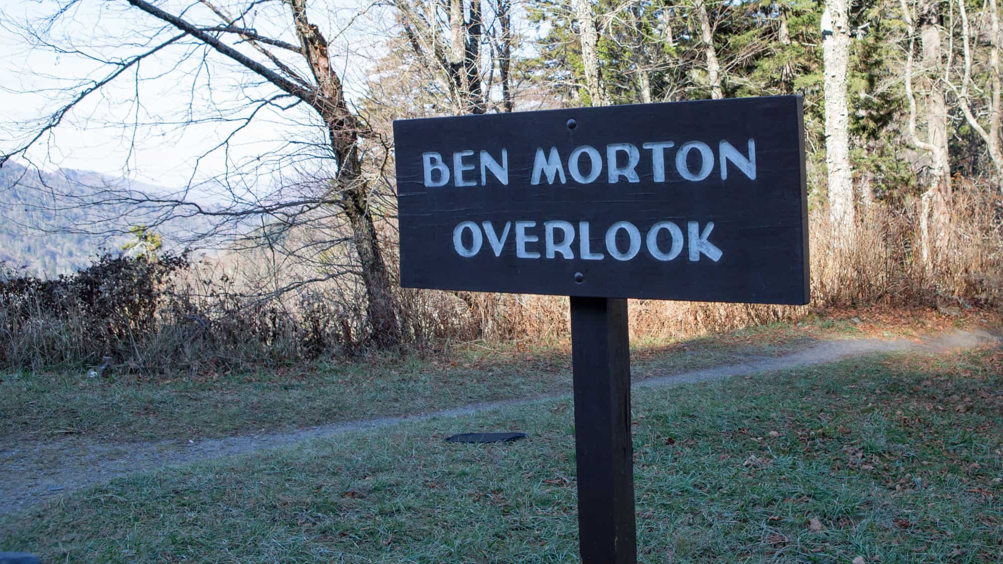 A wooden sign reading “Ben Morton Overlook” stands beside a forest trail, with bare trees and distant mountain views in the background.
