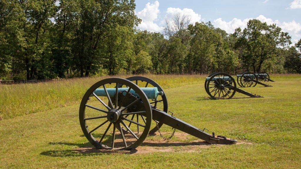 A line of vintage cannons is displayed across an open grassy field bordered by trees, symbolizing a preserved Civil War battlefield site.