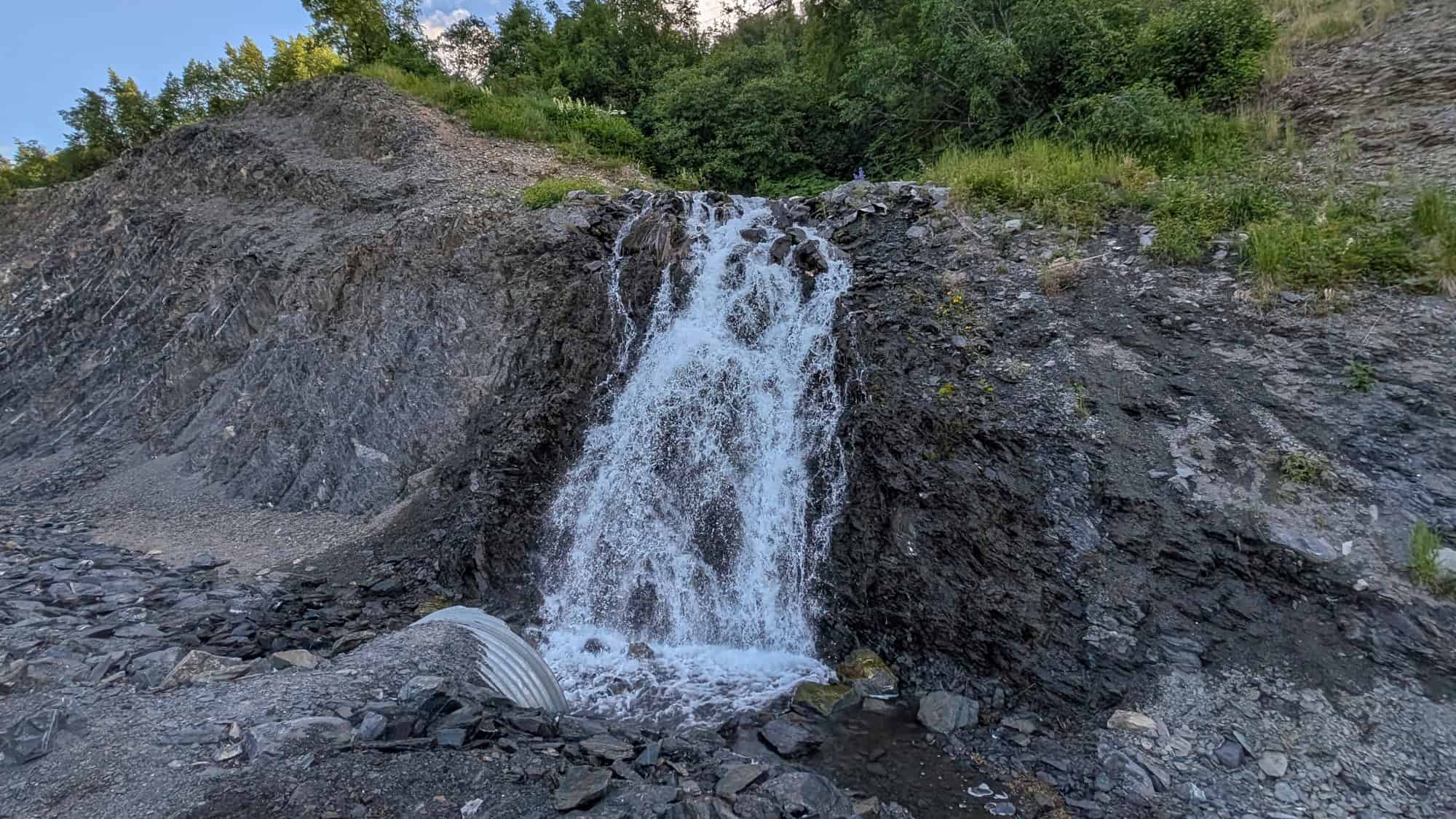 A small, powerful waterfall spills down dark slate rock beside a road, with greenery growing at the top of the slope.