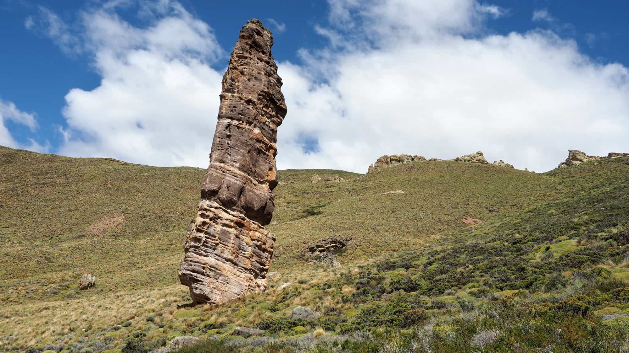 A towering, narrow rock spire juts out from a grassy hillside under a bright sky dotted with fluffy white clouds.