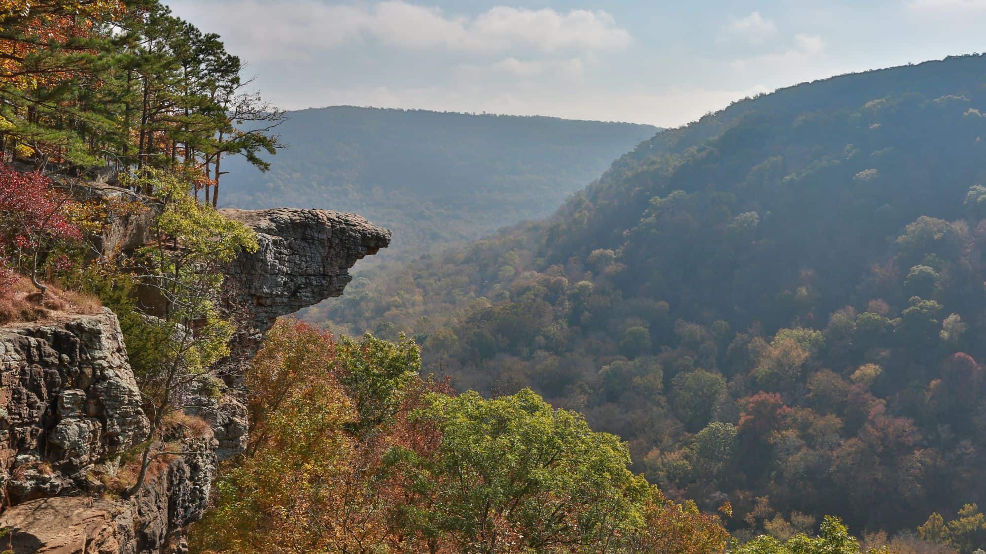 A famous rock outcrop extends dramatically over a forested valley with fall colors, framed by distant mountains and a hazy sky.