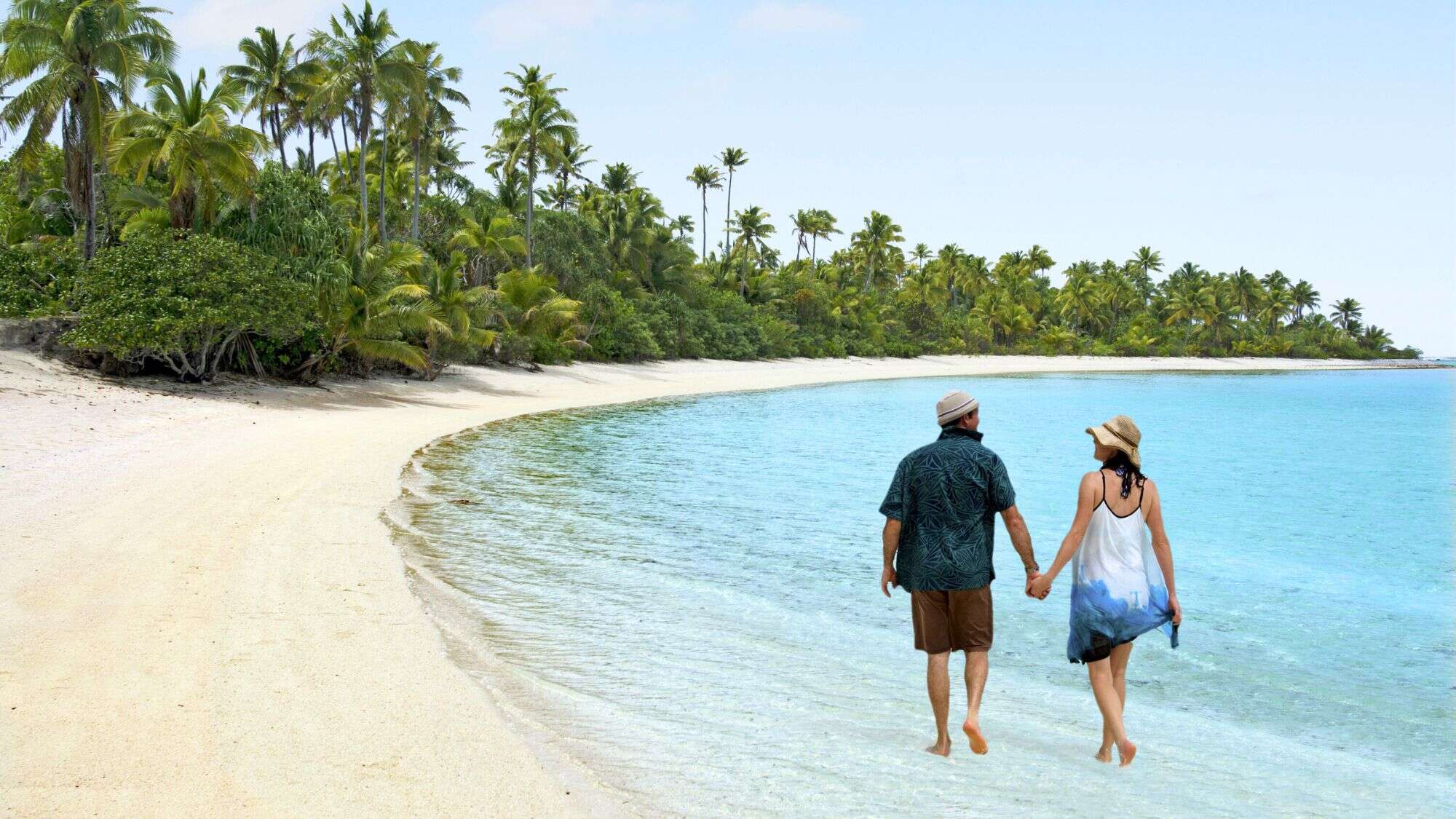A couple walks along a serene tropical beach with fine white sand and gently swaying palms, surrounded by clear turquoise water. The peaceful shoreline and lush greenery create a secluded island atmosphere.