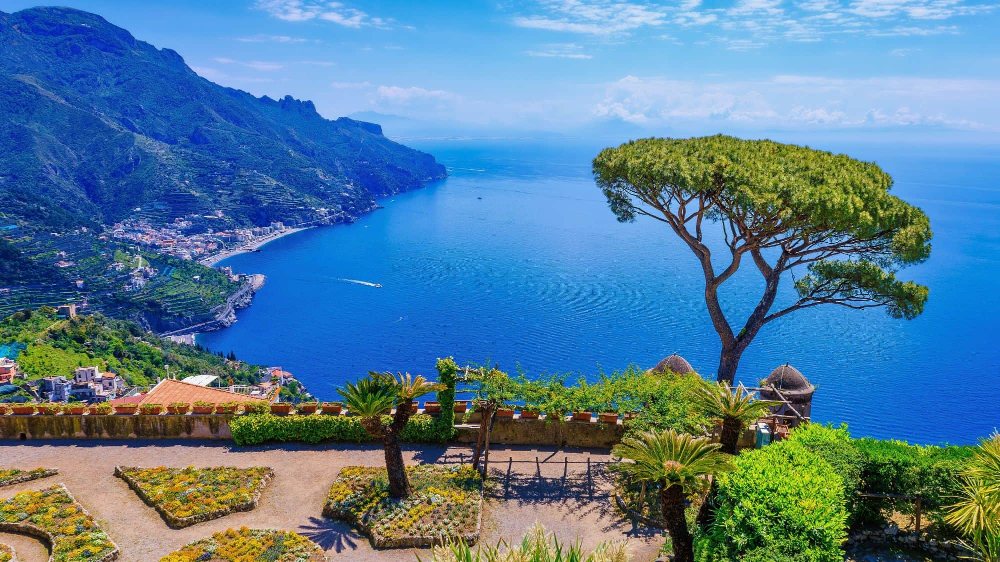 Colorful terraced gardens and a lone pine tree overlook the dazzling blue Tyrrhenian Sea, with hillside villages dotting the cliffs below. This view from Ravello captures the dramatic beauty of southern Italy’s coast.