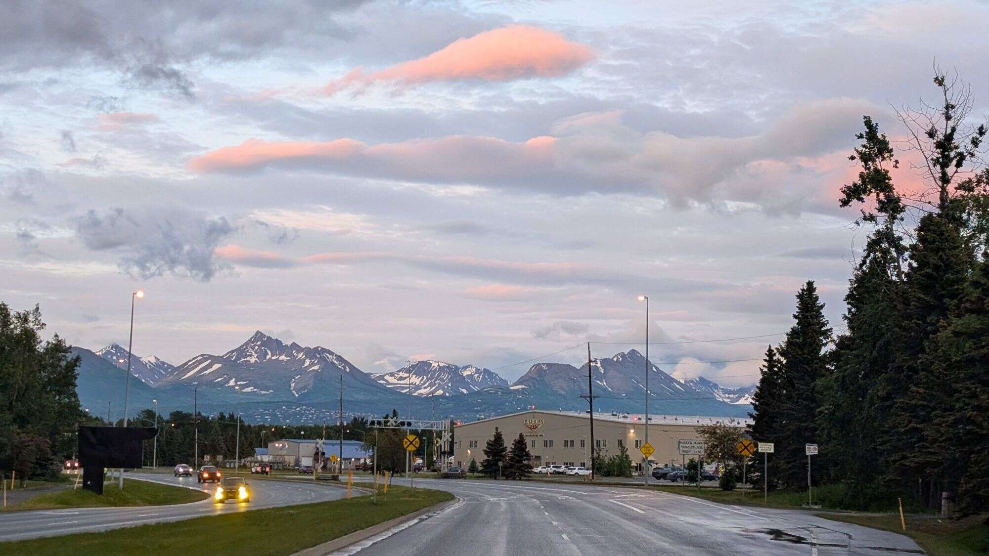Clouds tinged with soft pink hover above the Chugach Mountains as the road gently curves through Anchorage, backed by dramatic snow-capped peaks and an early evening glow.