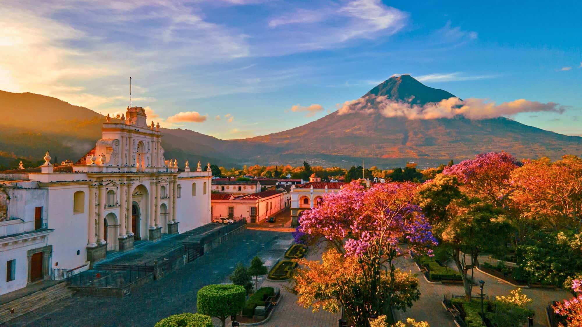 A historic white cathedral stands in soft evening light, with blooming trees in the foreground and a towering volcano rising dramatically in the background.