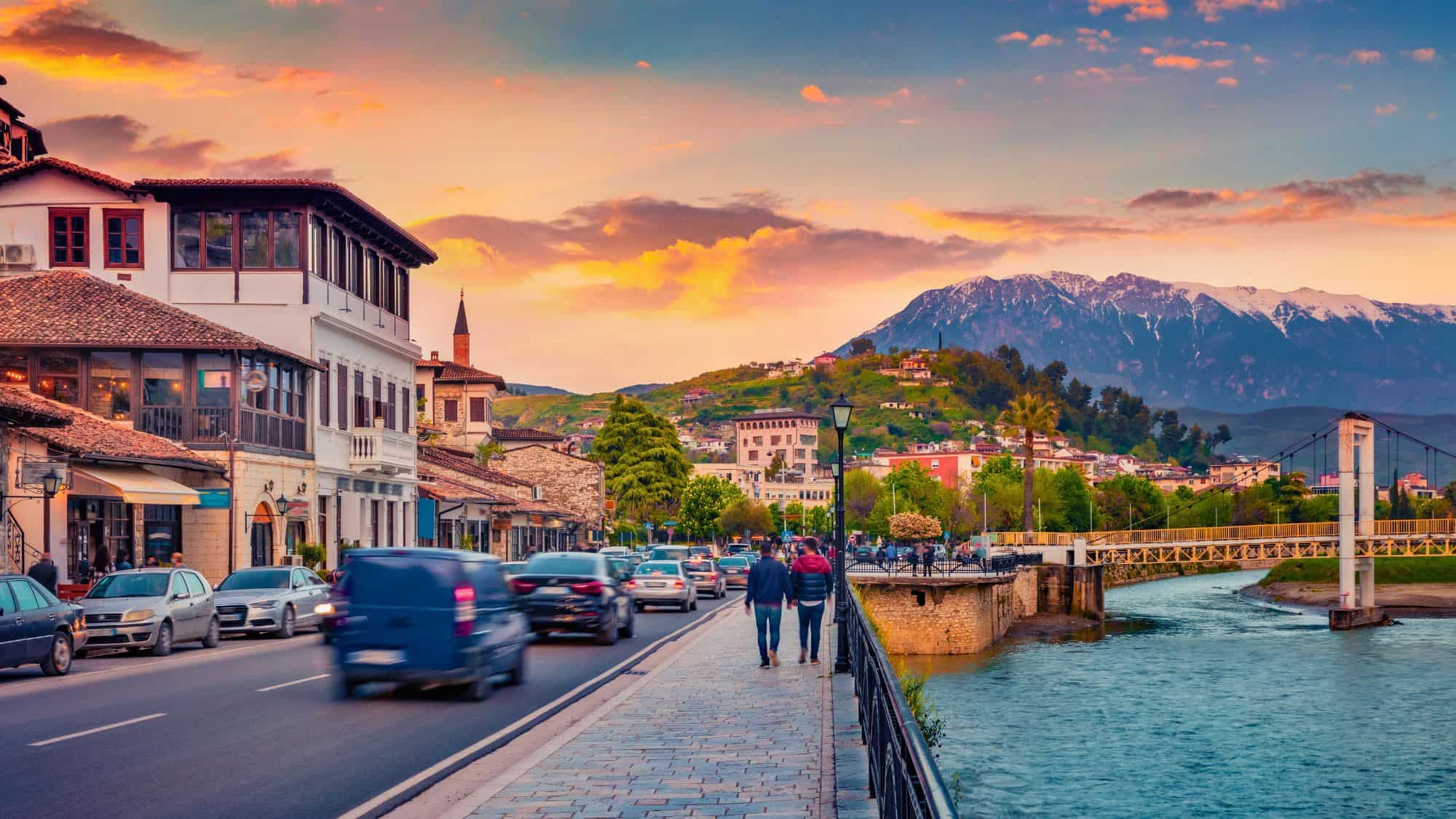 A cobblestone promenade runs alongside a lively street lined with traditional Ottoman-style buildings, set against a colorful sky and snow-capped mountains.