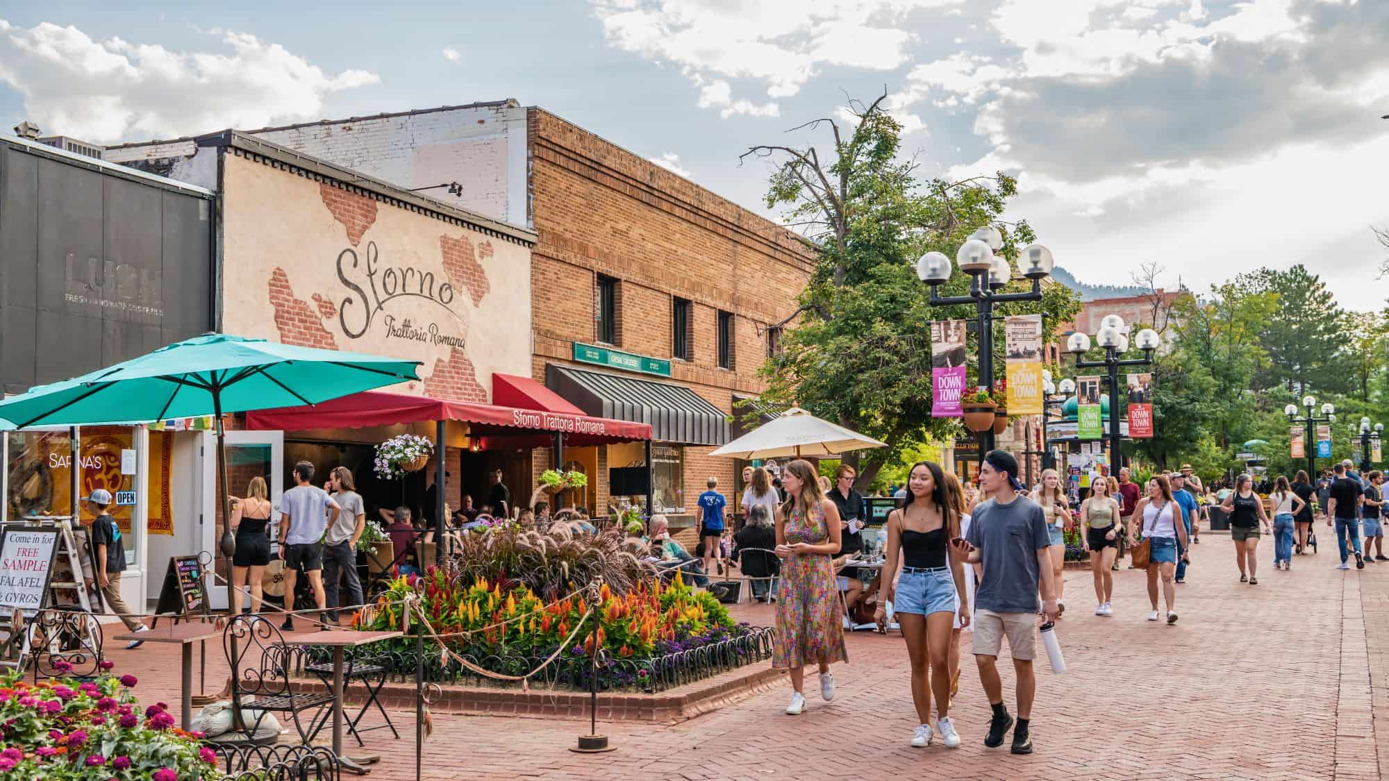 A lively downtown scene shows people strolling past restaurants, boutiques, and colorful banners along a brick-paved promenade lined with flowers and outdoor seating.