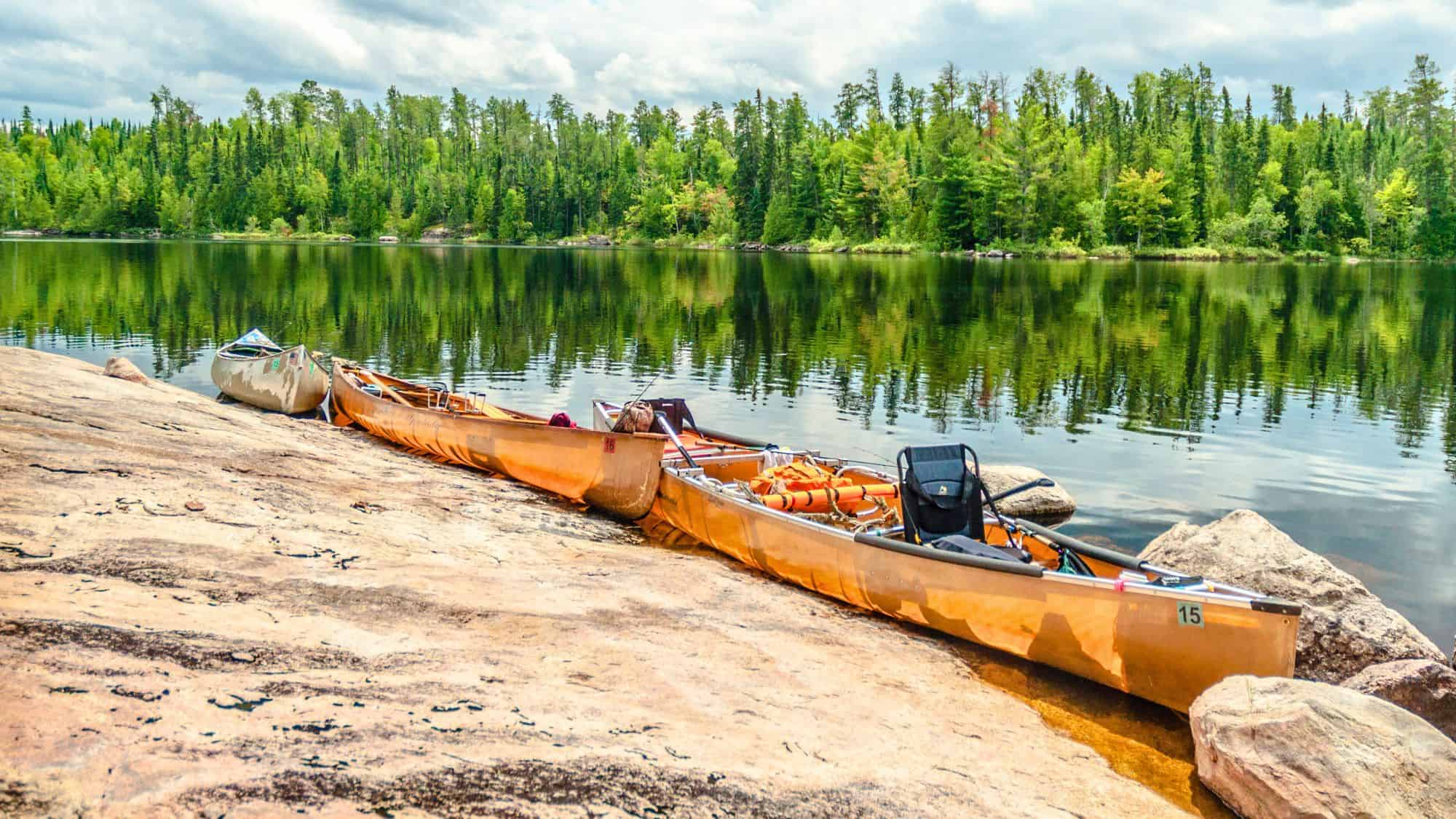 Three canoes rest on smooth rock beside a glassy lake, their reflections mirrored by a dense forest of green under a cloudy sky.