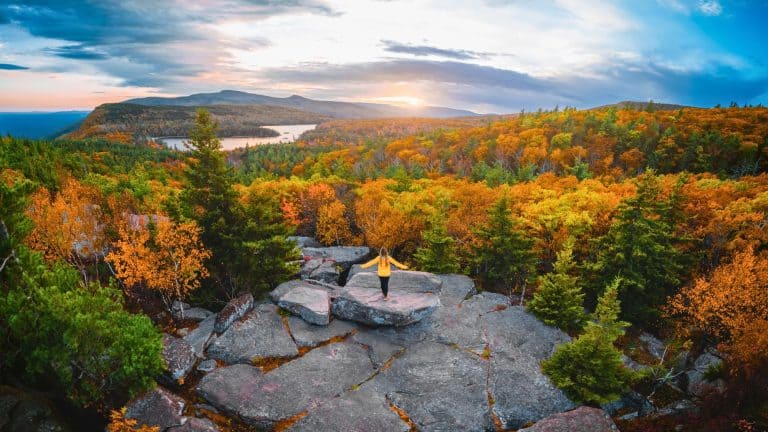 A woman stands on a rocky outcrop overlooking a vast expanse of vibrant autumn foliage, with lakes and distant mountains glowing under a dramatic sky.