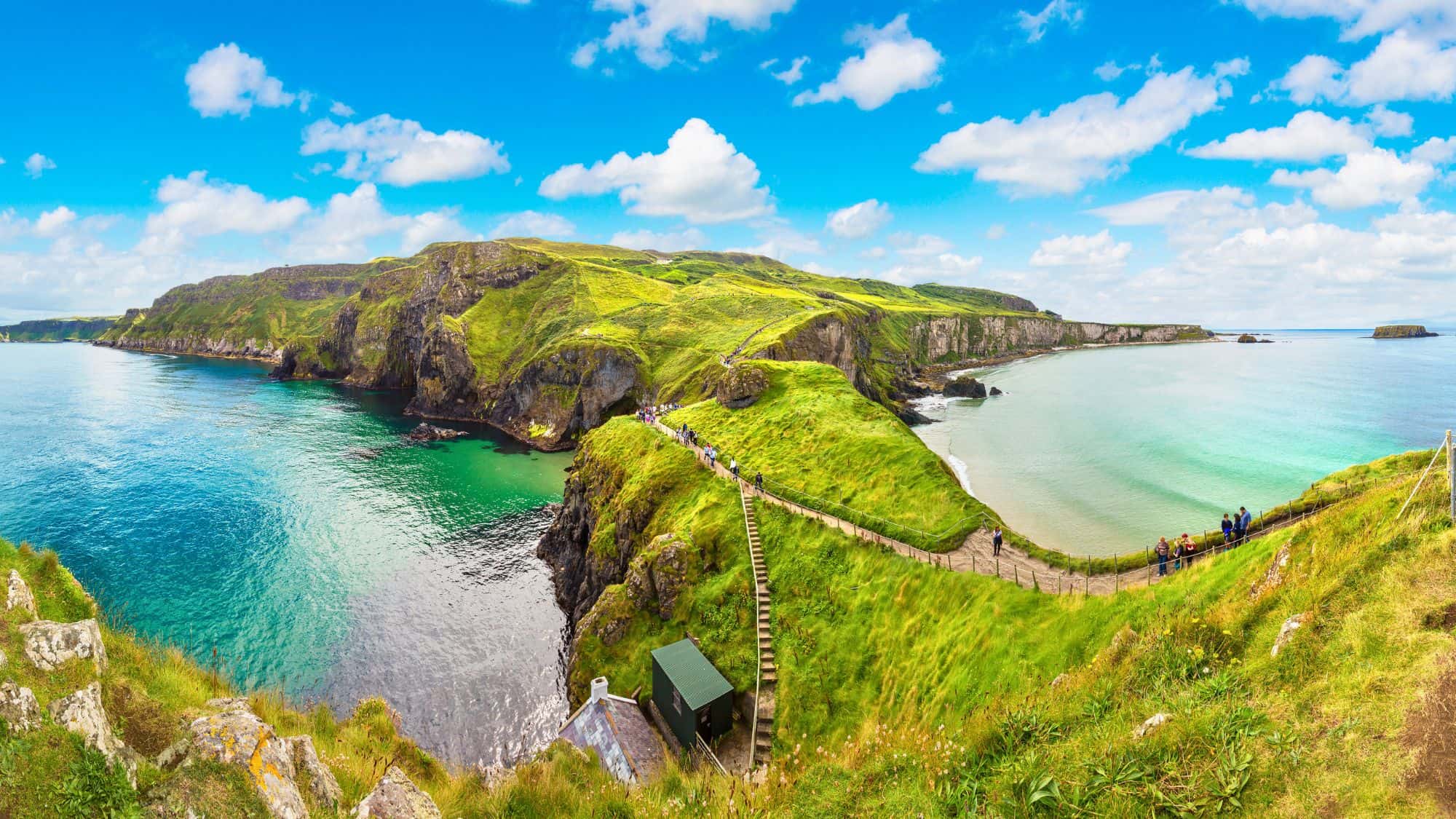 A narrow rope bridge connects the mainland to a rugged island surrounded by emerald cliffs and crystal-clear waters, with panoramic views of the Atlantic coastline.