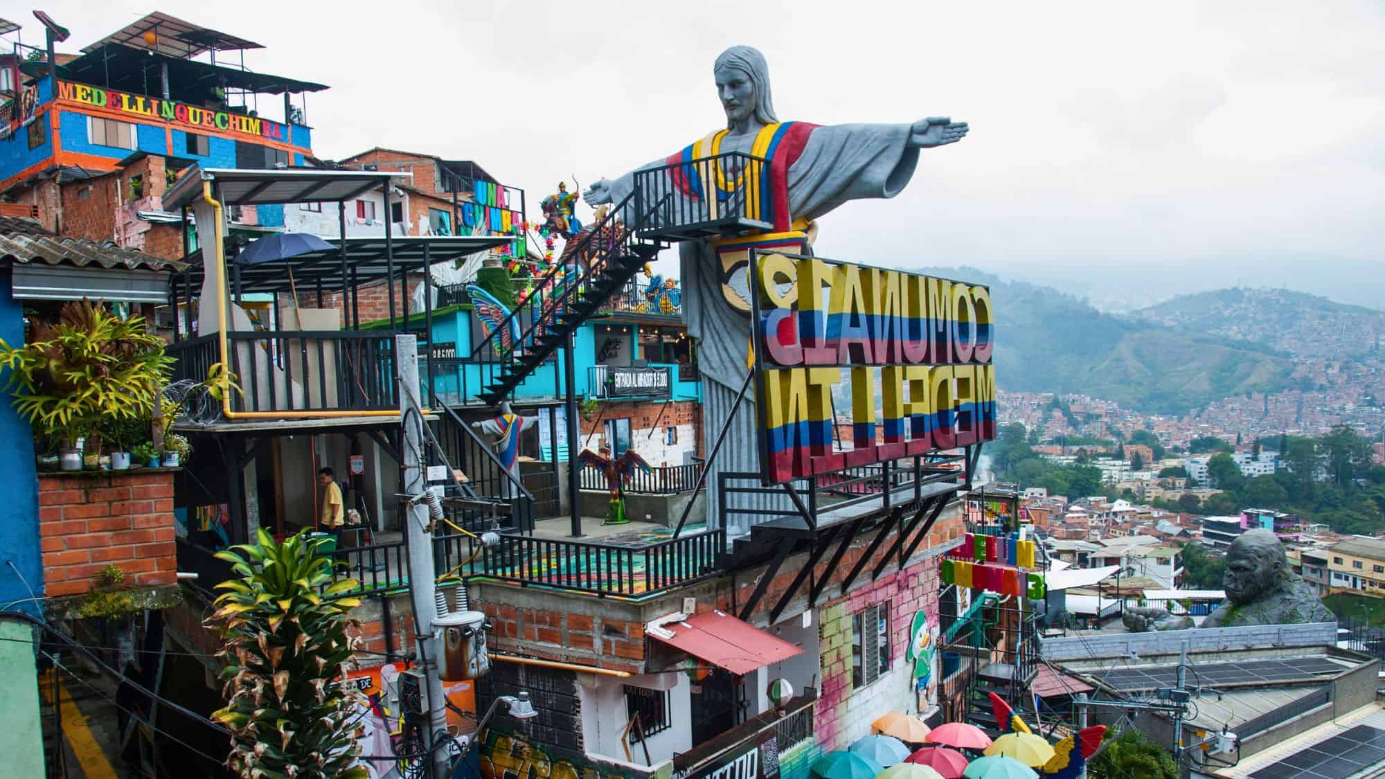 A hillside in Medellín bursts with color from murals, sculptures, and stacked houses, crowned by a large Christ statue draped in Colombia’s flag colors.