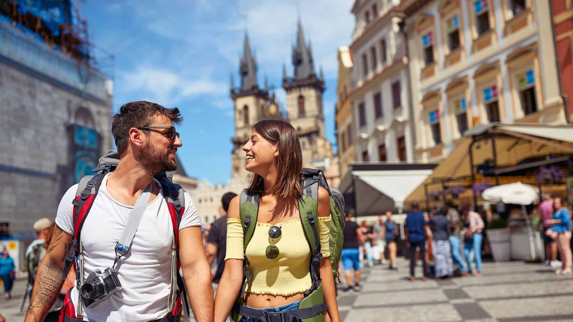 A couple walks hand in hand through a bustling square lined with ornate buildings, while Gothic spires of a historic church rise in the background.