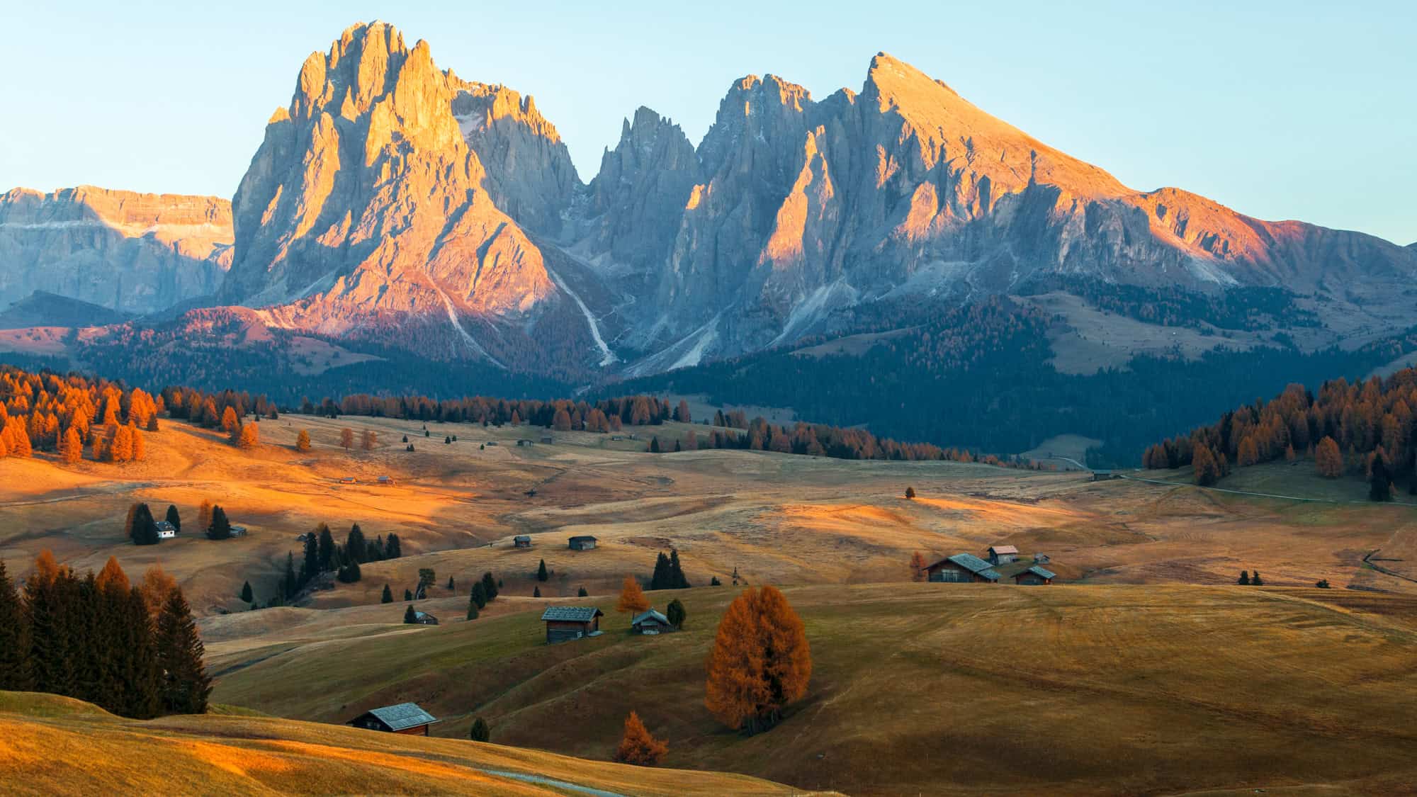 Golden morning light washes over gentle meadows and scattered wooden huts, while the jagged peaks of the Dolomites rise sharply in the background.