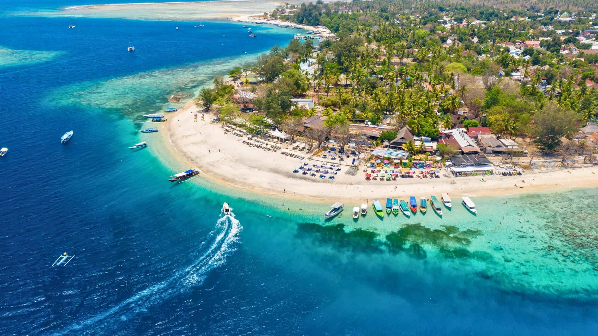 Aerial view of a white-sand beach lined with colorful boats and palm trees, curving into bright blue waters along a lush island coast.