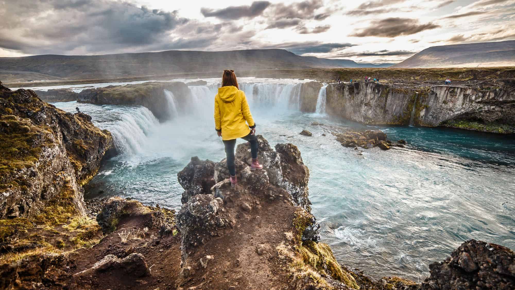 A person stands near the edge of dramatic cliffs, watching the powerful cascade of glacier-fed water.