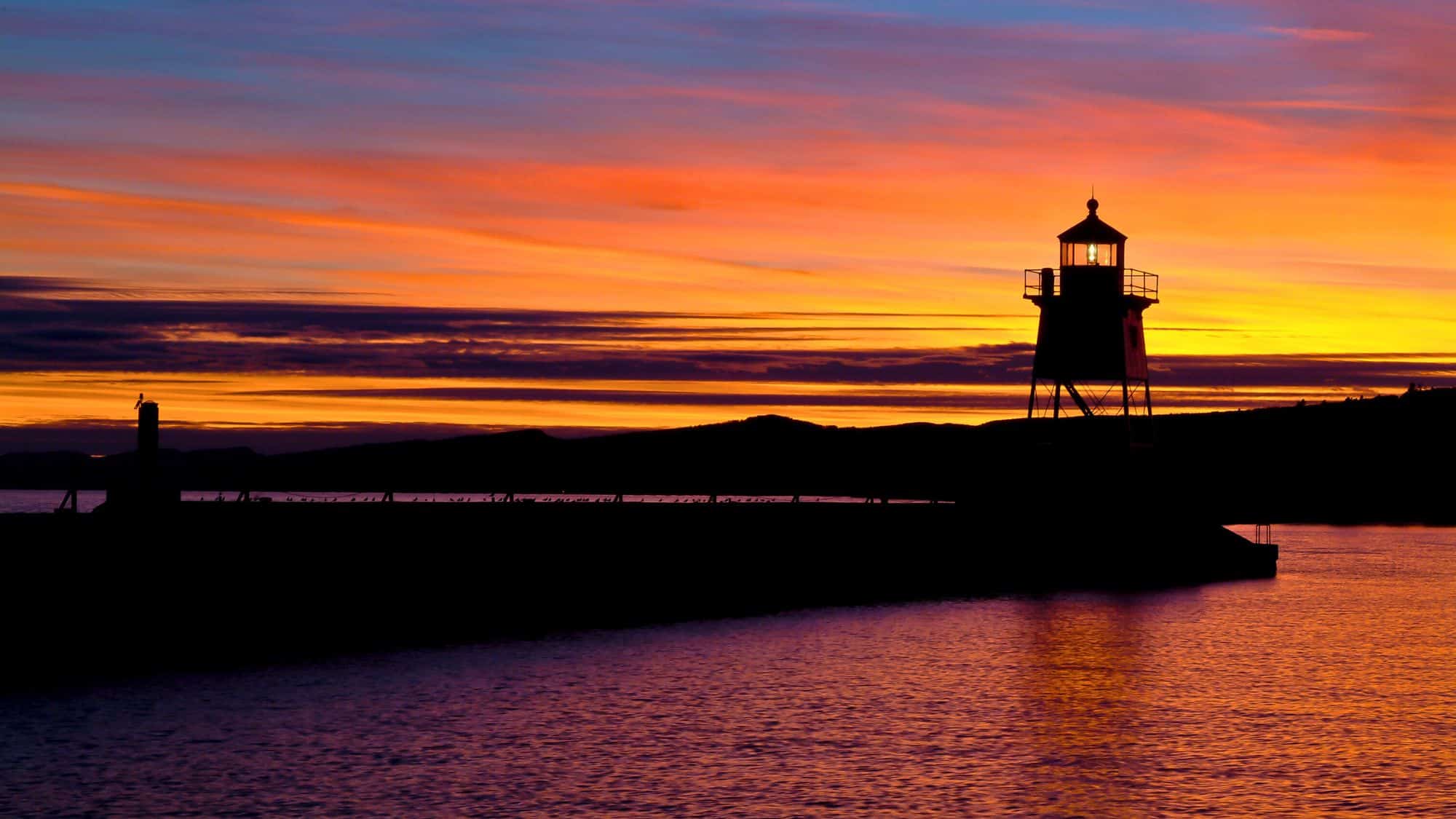 A glowing sky drenched in rich purples, oranges, and golds silhouettes the historic Grand Marais lighthouse along Lake Superior’s tranquil shoreline.