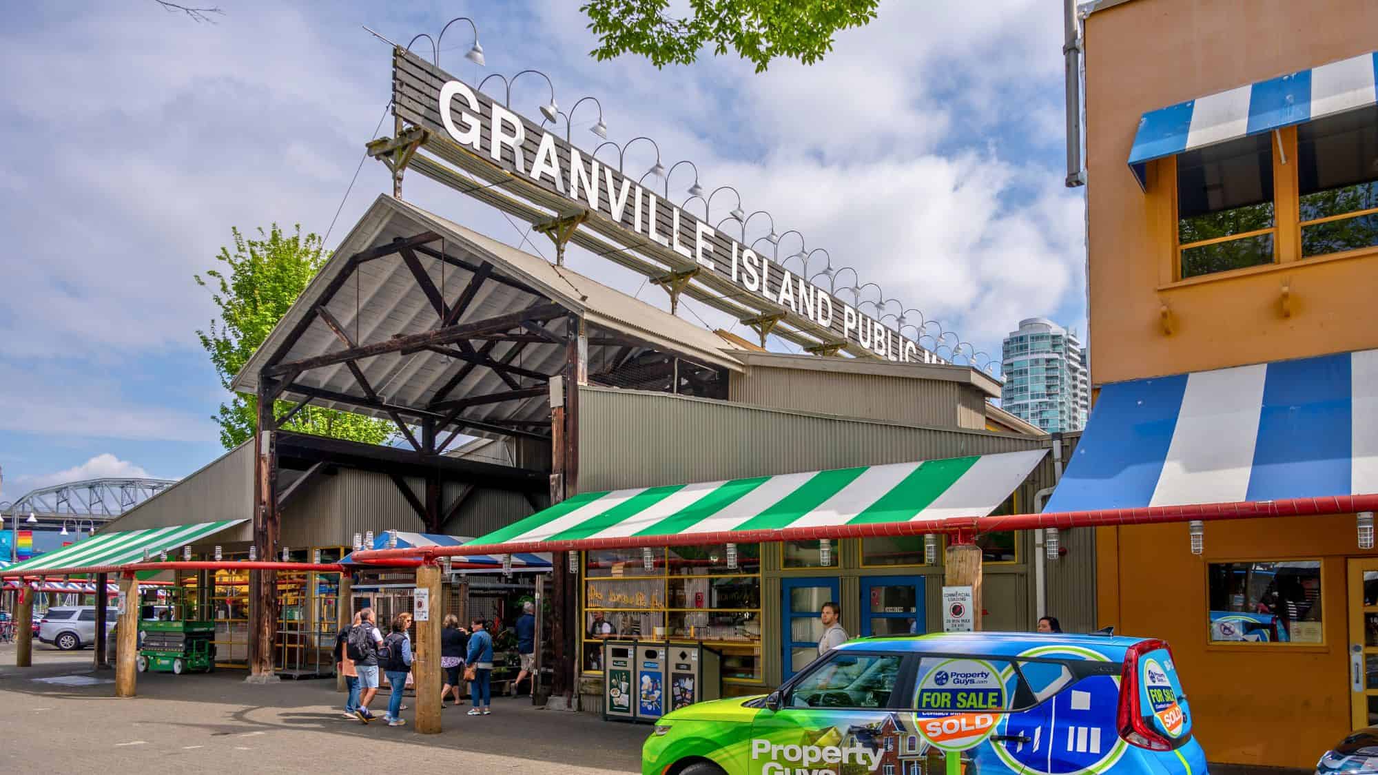The entrance to this bustling market is framed by green and white striped awnings, with shoppers exploring food and craft stalls beneath the bold sign overhead.