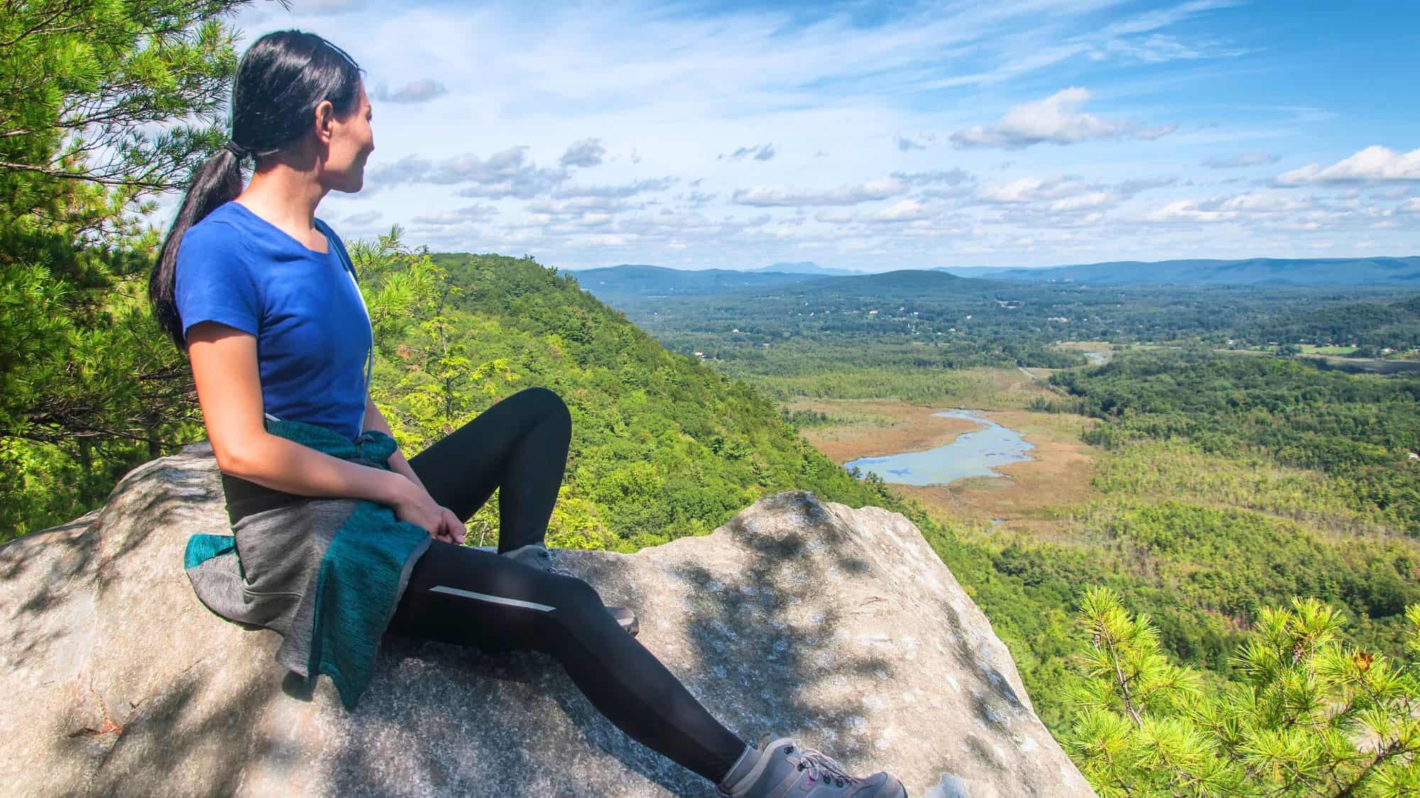 A woman sits on a sunlit boulder, taking in a sweeping view of forested hills, winding waterways, and distant ridgelines under a clear blue sky.