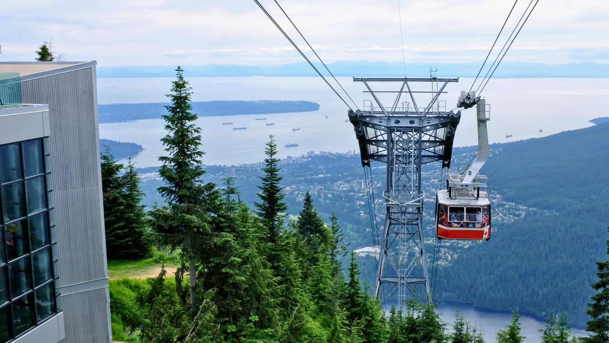 A cable car ascends above dense evergreen forest, offering expansive views of Vancouver, the bay, and distant ships dotting the blue horizon.