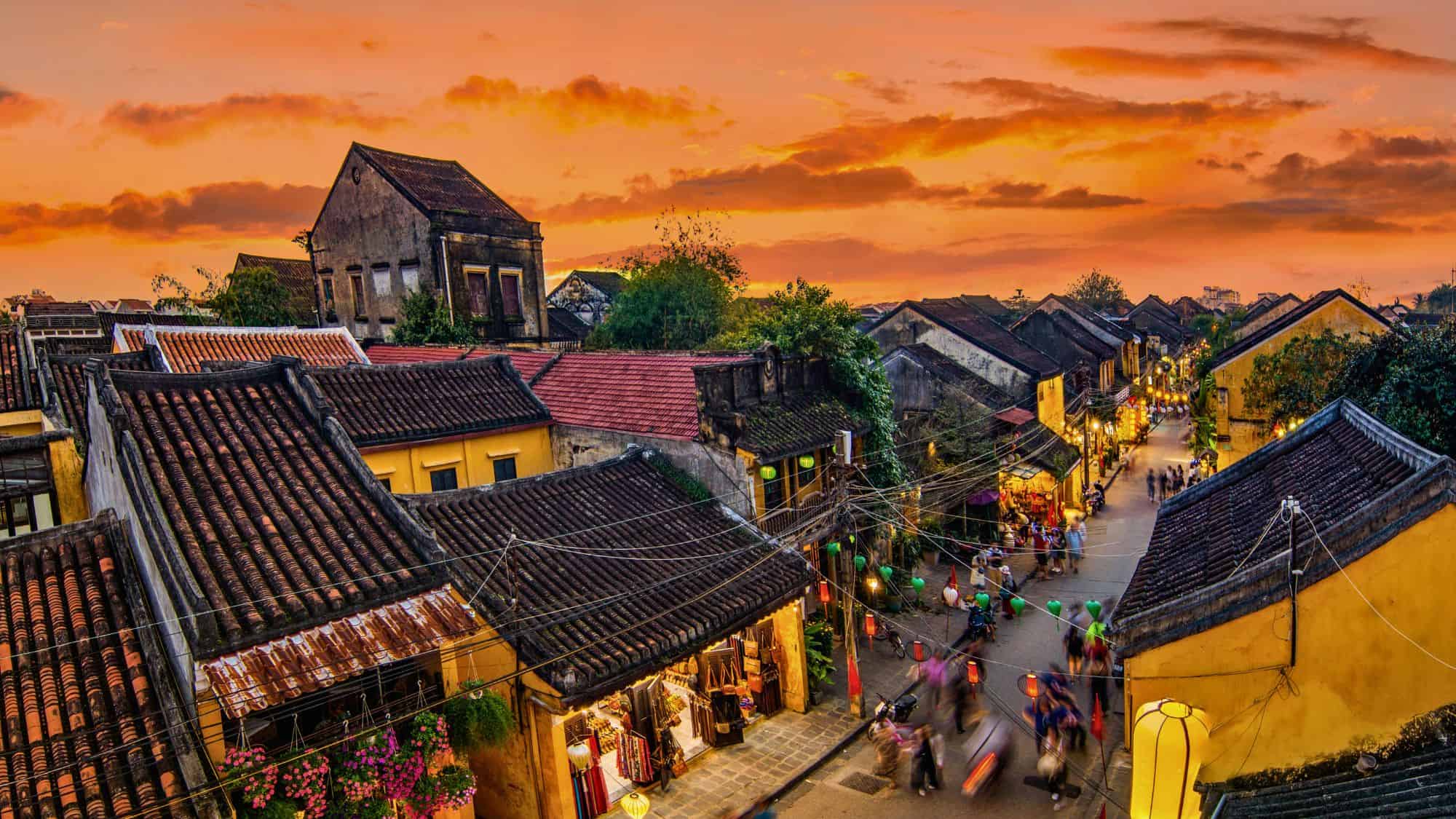 Lantern-lit streets wind between yellow-walled shops and tiled roofs as the sky glows orange, capturing the lively charm of Vietnam’s historic district.