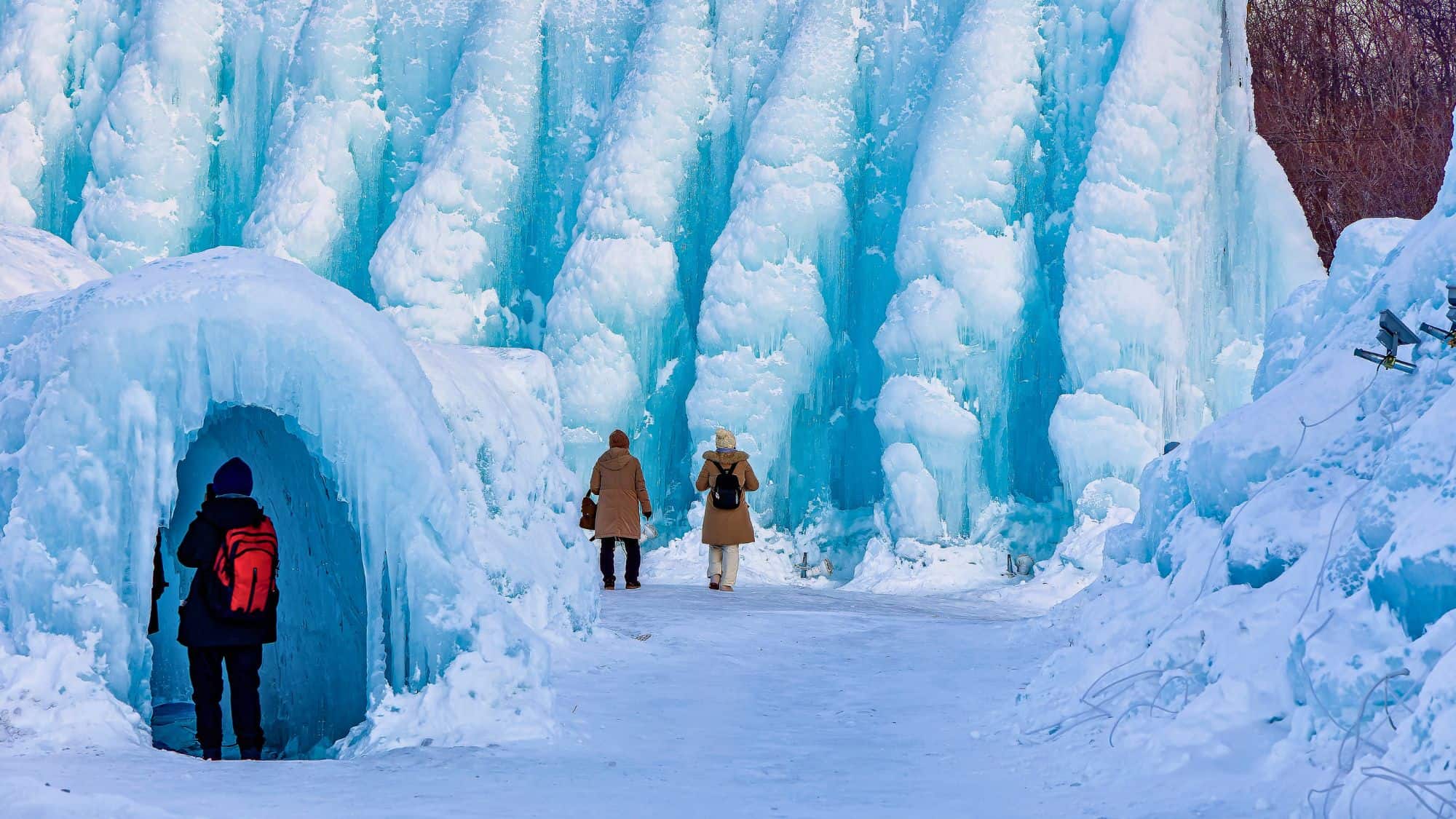 Massive icy formations tower over bundled-up visitors exploring an otherworldly winter scene with glowing blue hues and frozen tunnels.