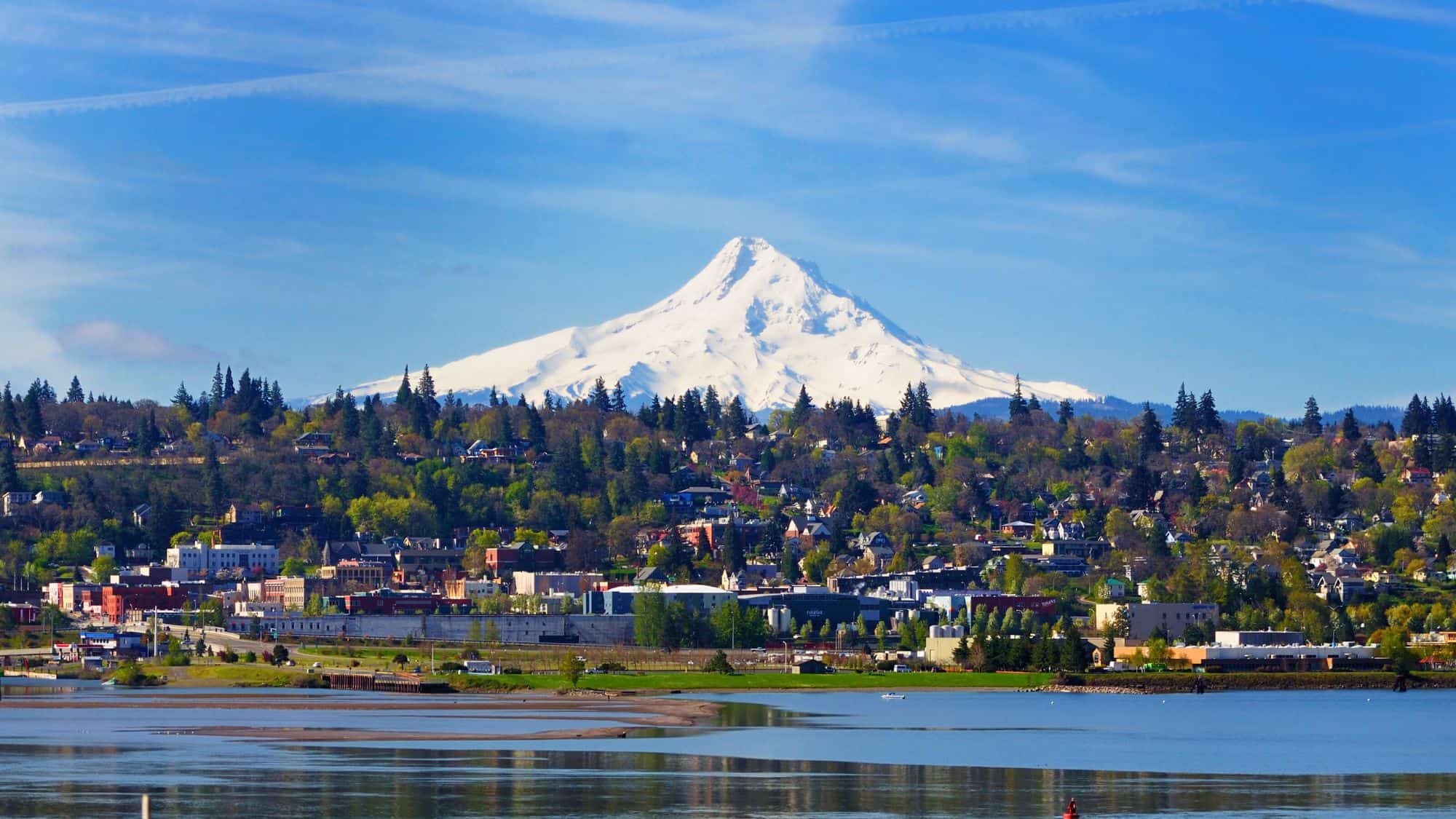 Snow-capped Mount Hood rises dramatically behind the town of Hood River, Oregon, where houses and trees fill the foreground under a bright blue sky.