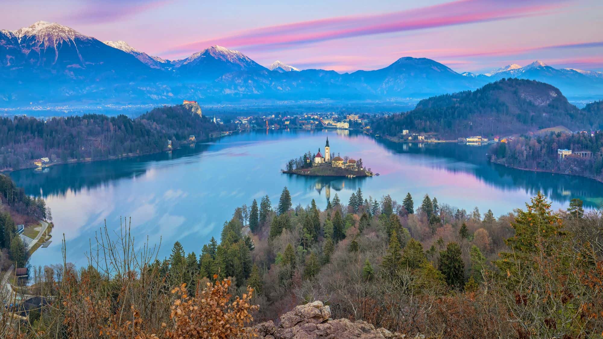 A tiny church-topped island floats on a still alpine lake surrounded by snow-capped mountains, all under a soft purple and pink sky.