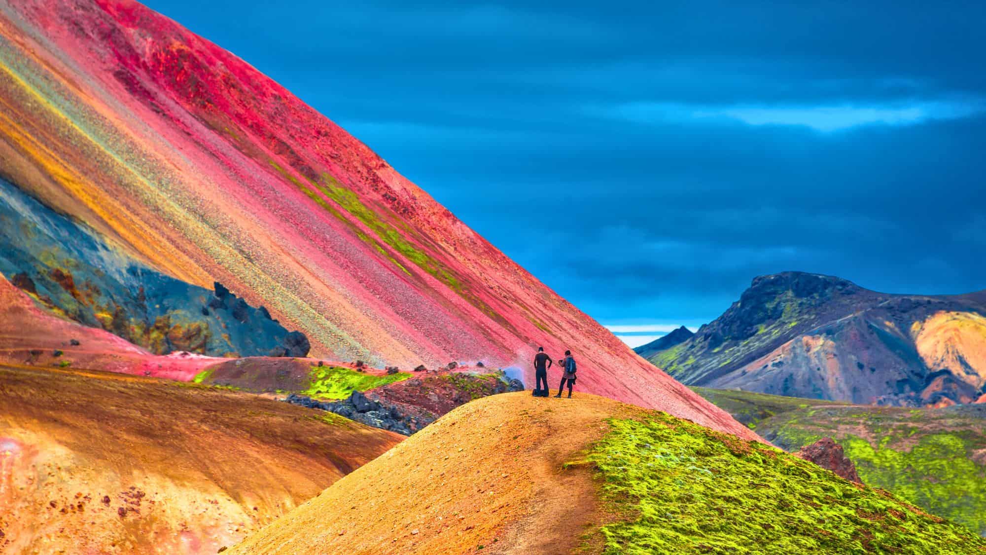 Two hikers stand before a surreal mountain slope striped in vivid reds, greens, and golds beneath a deep blue sky in a volcanic landscape.