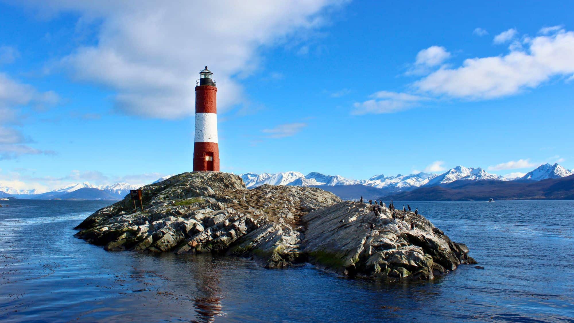 This red-and-white striped lighthouse stands on a rocky outcrop in the Beagle Channel, surrounded by cold waters and distant snow-capped mountains. It's a windswept landmark near the southern tip of South America.