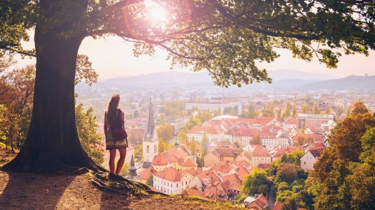 A woman gazes over the charming old town of Ljubljana, Slovenia, where pastel-colored rooftops, church spires, and leafy trees stretch into the hazy hills.