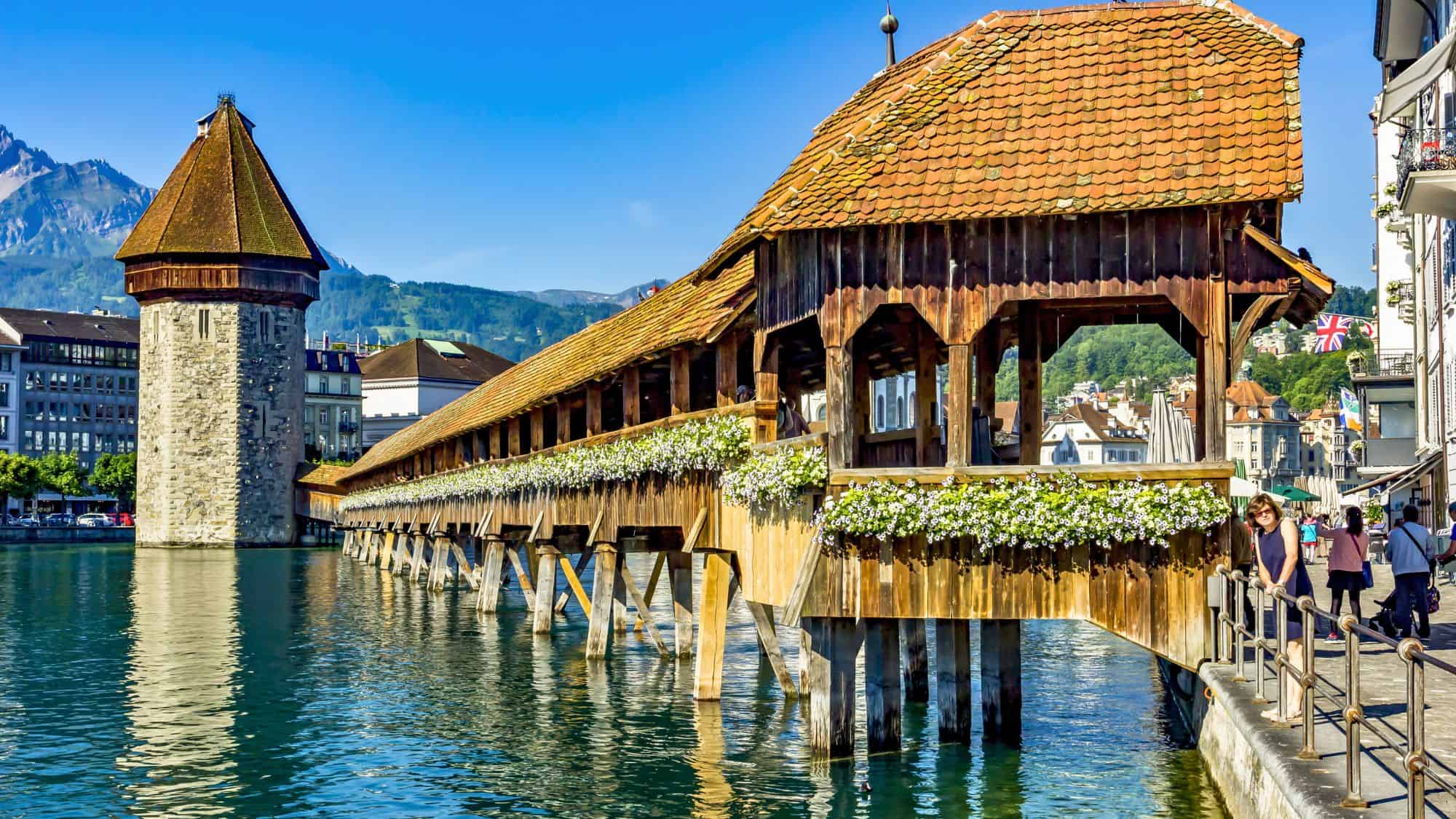 A historic covered wooden footbridge stretches across the Reuss River, adorned with vibrant flower boxes and backed by alpine peaks in Lucerne, Switzerland.