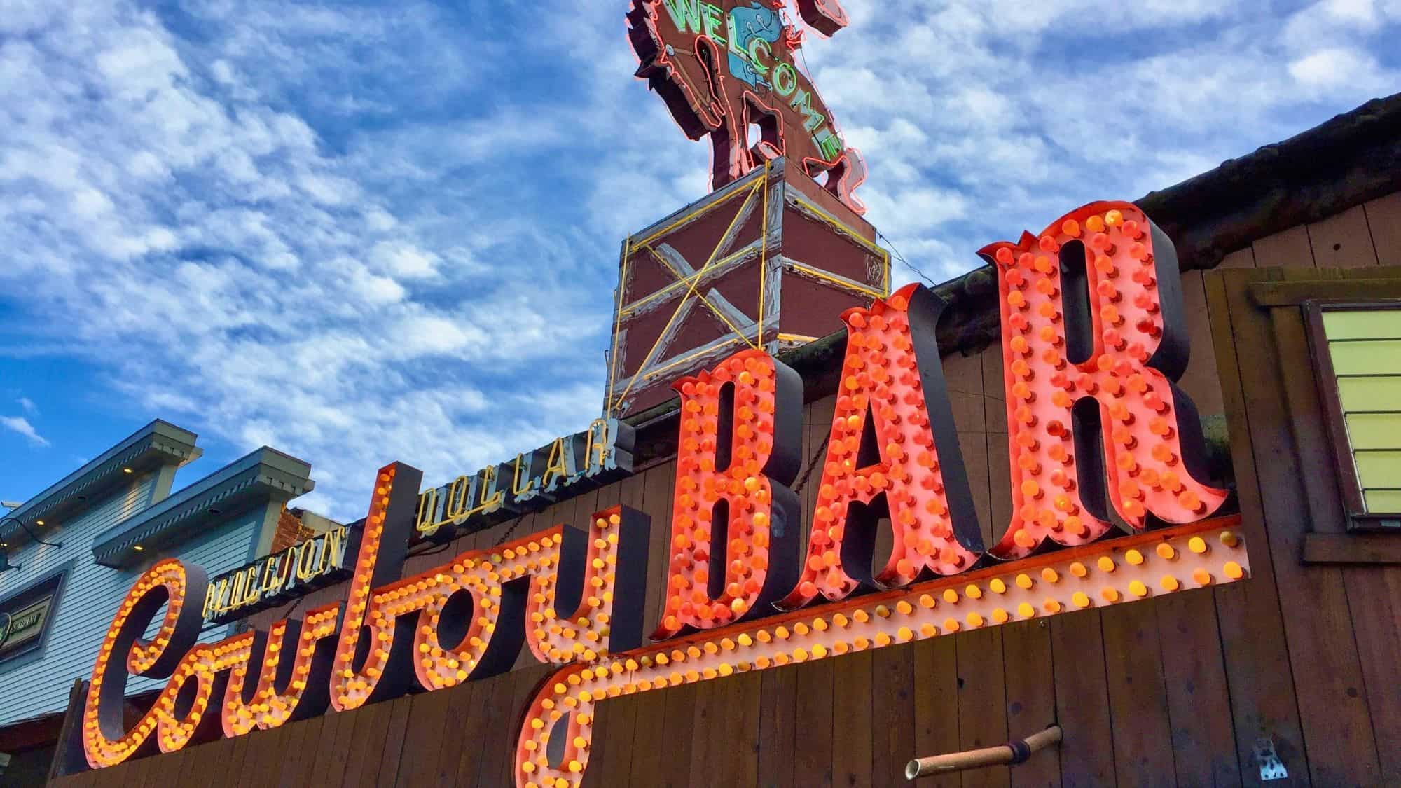 A classic Western-style bar sign lights up with bold retro bulbs under a bright blue sky, topped by a bucking cowboy silhouette.