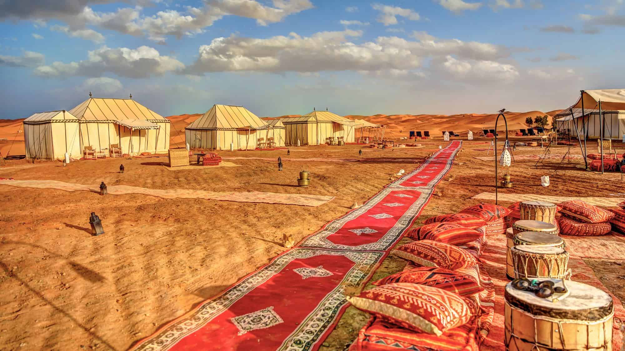 A Moroccan desert camp features elegant tents and patterned rugs stretched across orange sands, with drums and cushions set for sunset relaxation.