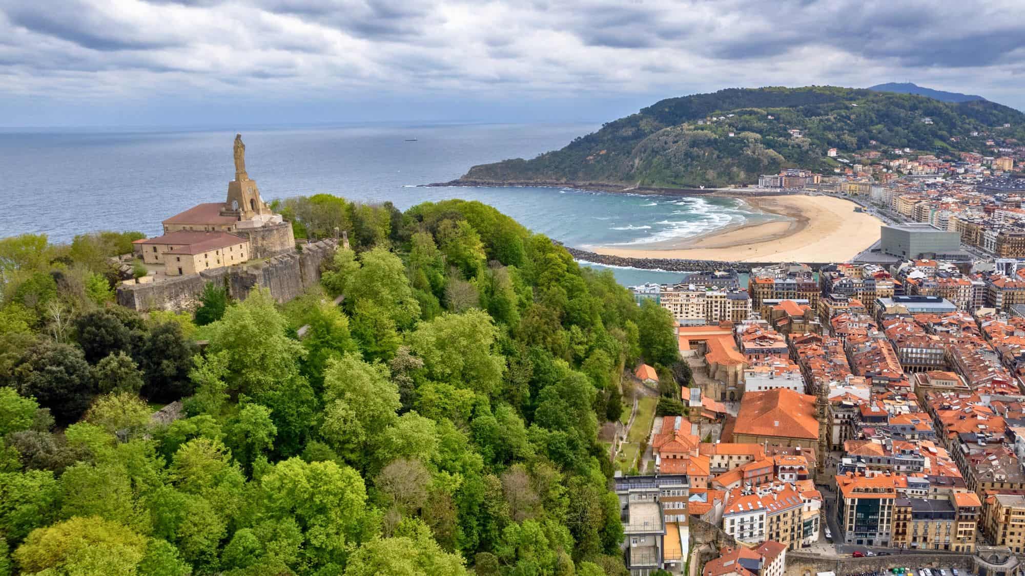 A lush green hill with a statue-topped fortress looks over San Sebastián's coastline and historic city center, where red-tiled rooftops meet the curving beach.