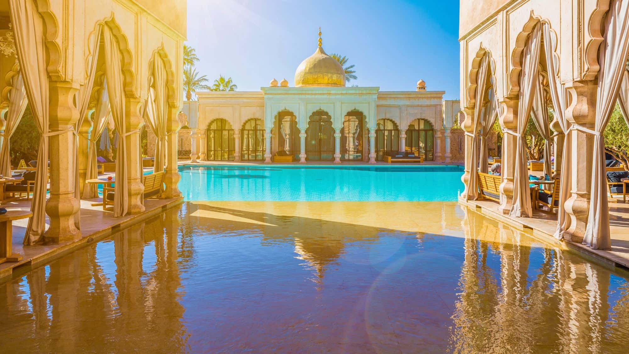 Golden arches and an opulent dome reflect in a sun-drenched pool, framed by shaded lounging areas and palm trees.