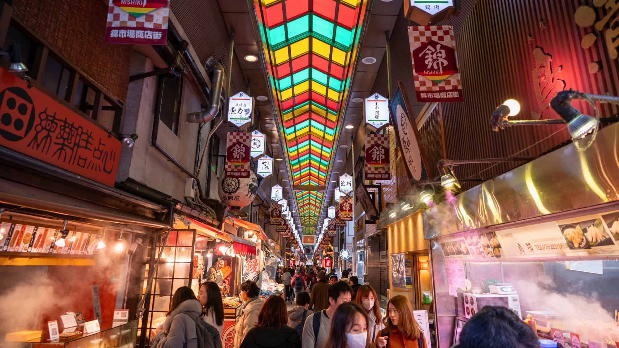 Crowds browse through a covered shopping street lined with steaming food stalls, lanterns, and a stained-glass ceiling glowing in red, yellow, and green tones.