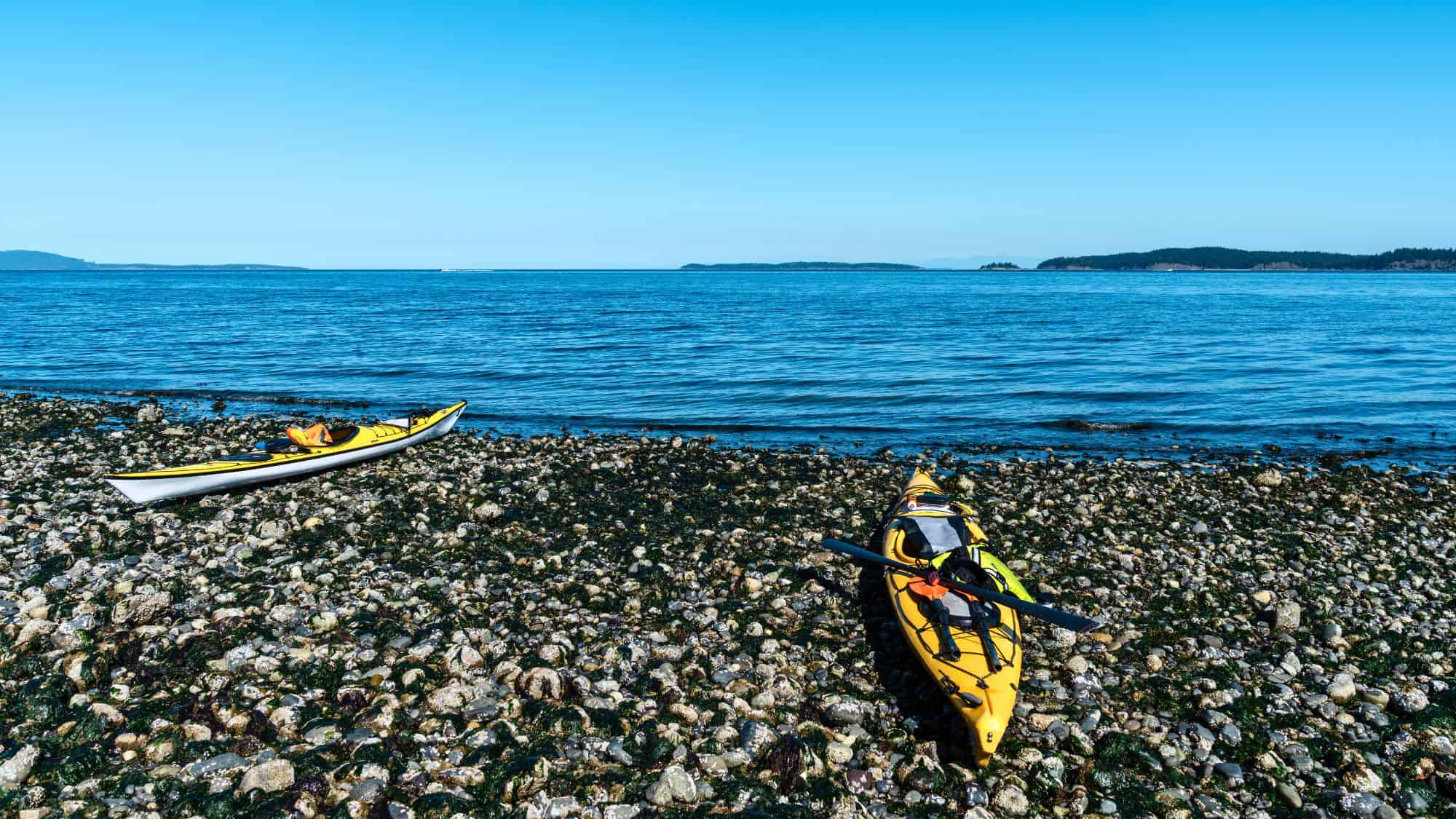 Two kayaks rest on a rugged, seaweed-strewn shoreline beside calm, open water under a bright sky, suggesting a quiet coastal stopover.