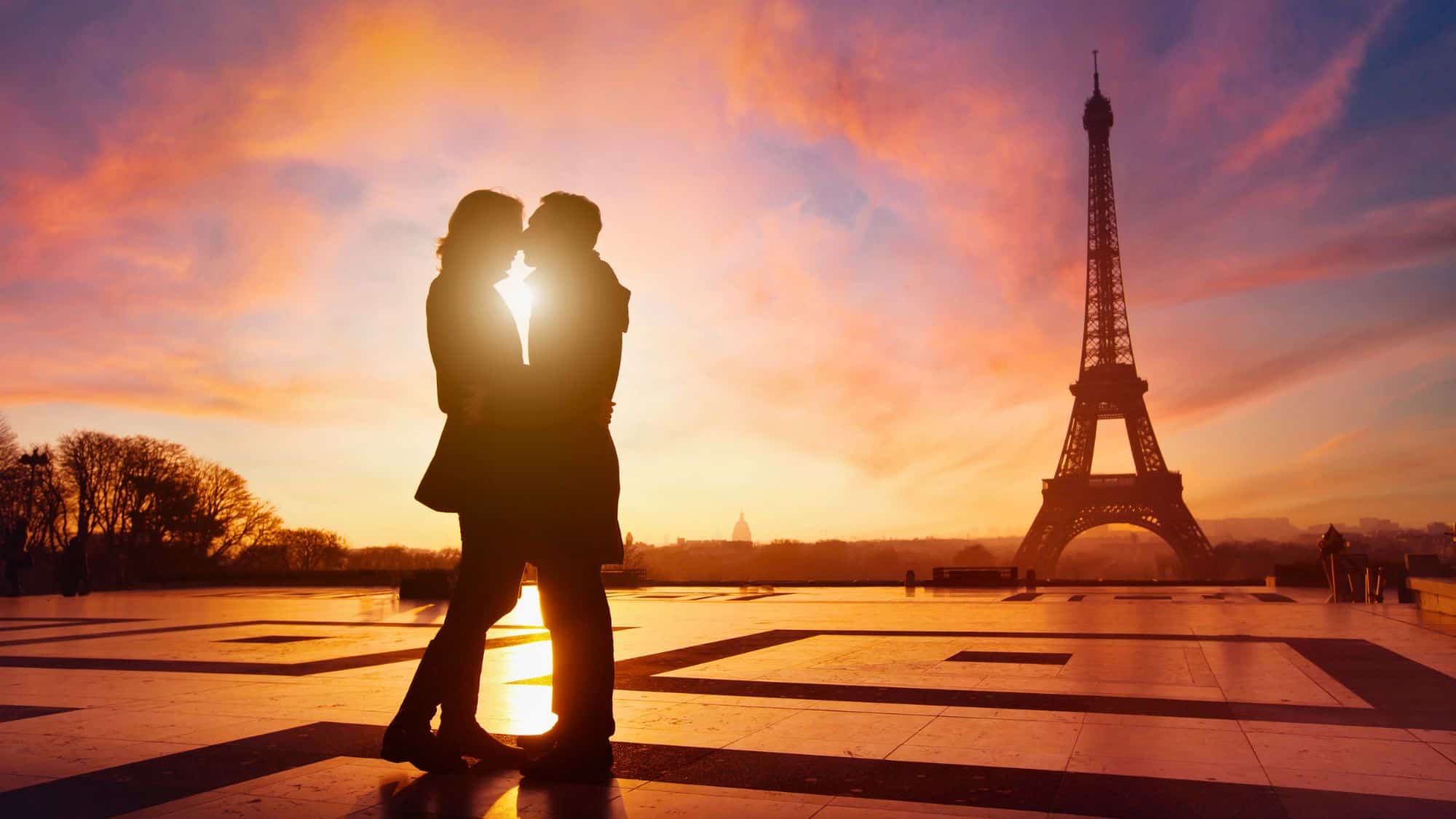 A silhouetted couple shares a moment in front of the Eiffel Tower at dusk, with the sky painted in warm shades of orange and lavender.