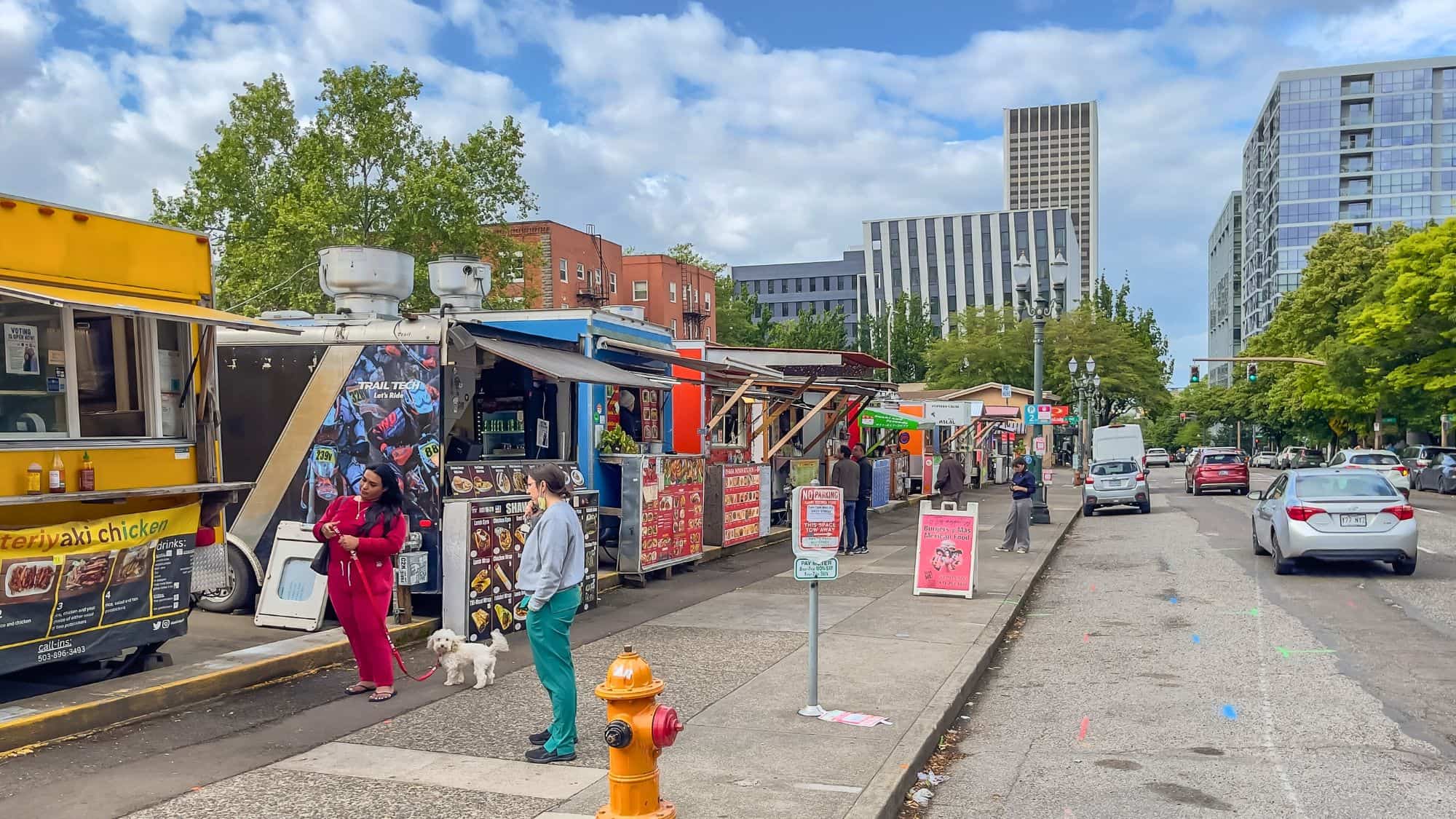 A row of colorful food trucks offers a variety of global street eats, with customers waiting and chatting under a mix of trees and urban high-rises.