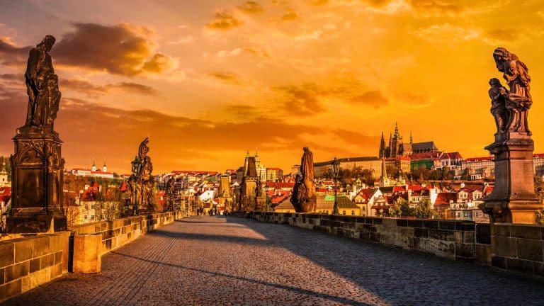 Statues line the historic Charles Bridge in Prague, glowing beneath a dramatic orange sky as the city’s skyline and castle rise in the distance.