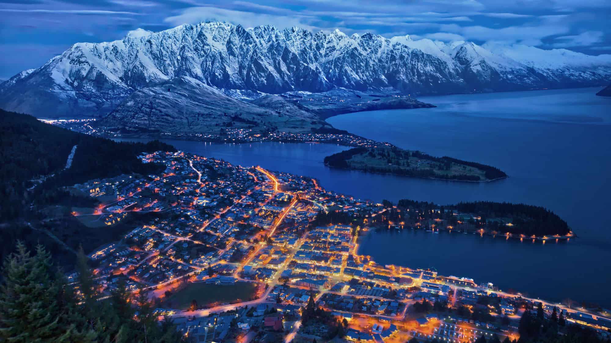 This twilight view of Queenstown, New Zealand captures the town’s glowing lights beneath snow-covered peaks and the sweeping curve of Lake Wakatipu.