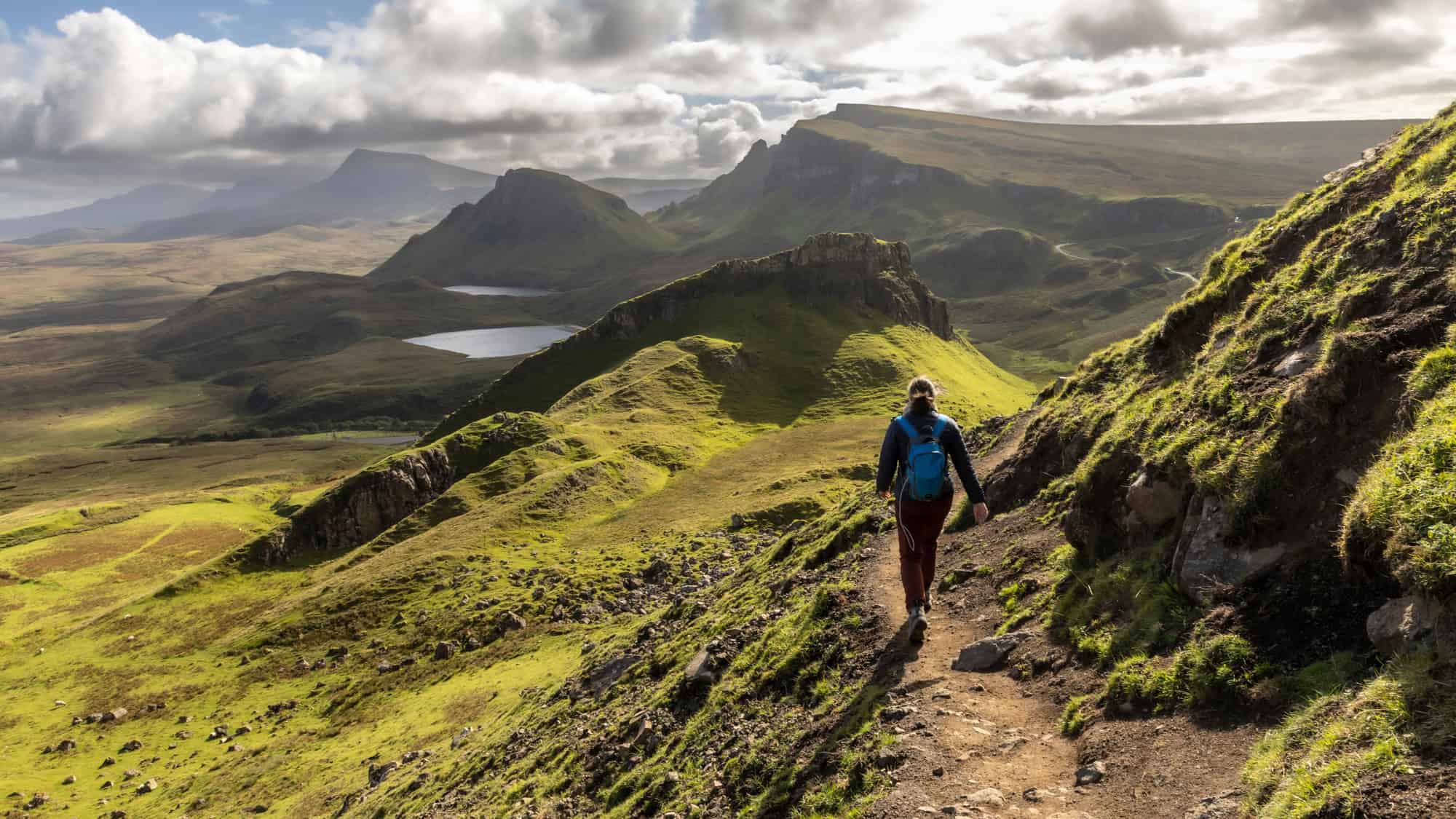 A lone hiker walks along a rugged trail carved into rolling green hills with dramatic rock formations and scattered lochs stretching into the mist.