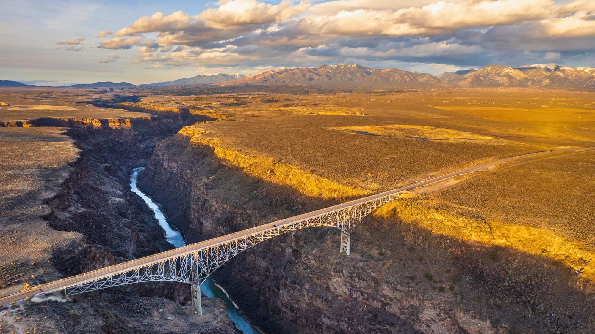 An impressive steel bridge spans the deep chasm of the Rio Grande Gorge in New Mexico, set against a golden high desert landscape with mountains in the distance.