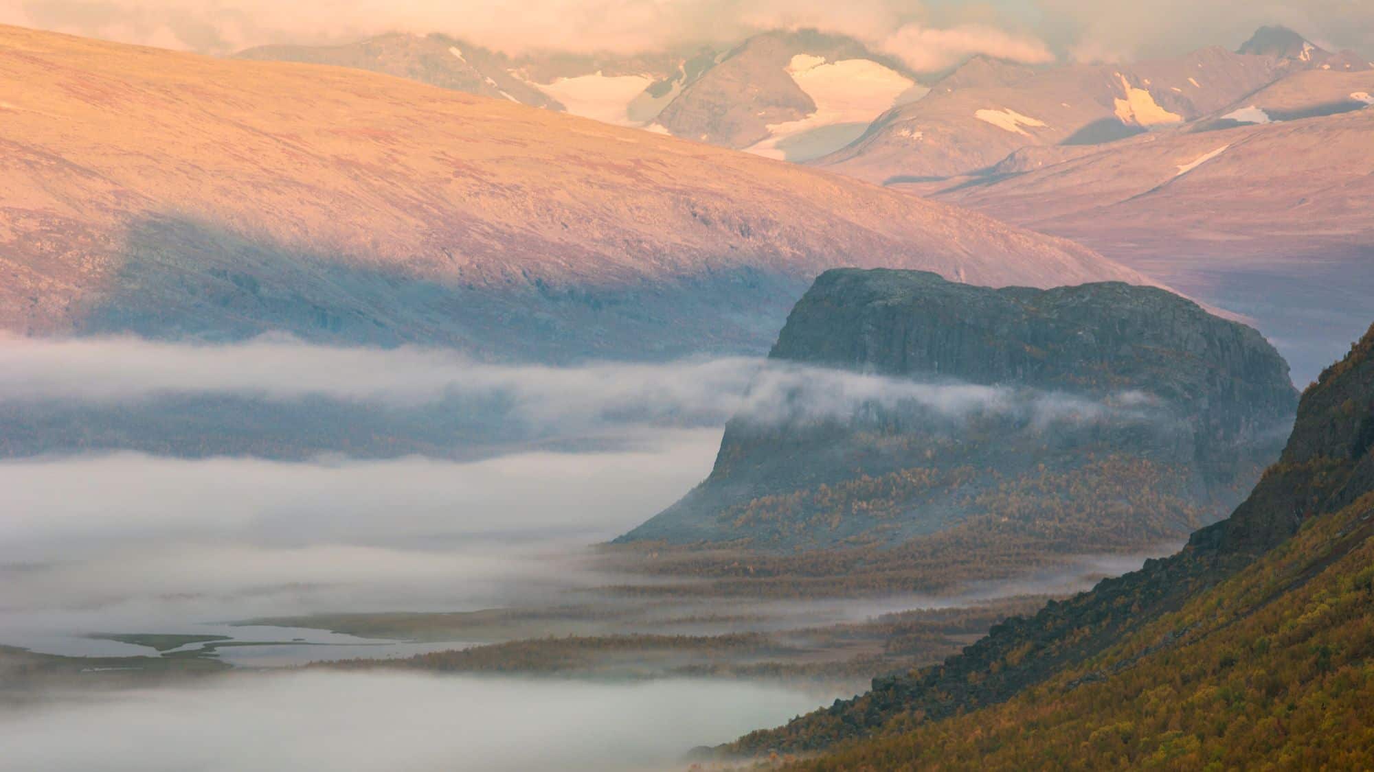 Thick morning fog drapes the wide tundra valley and wraps around steep cliffs, with glacier-topped mountains glowing in soft light above.