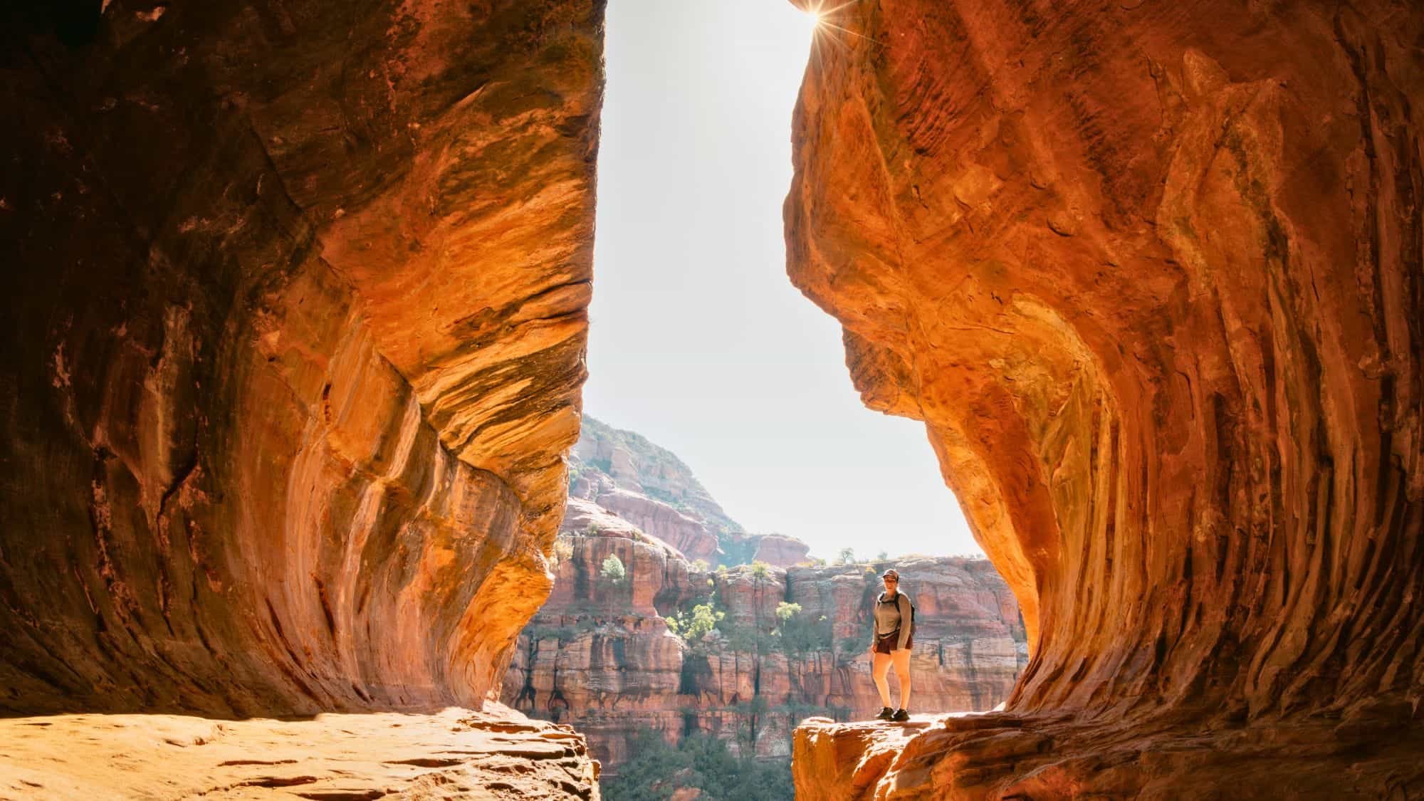 Framed by smooth red rock walls, this secluded cave near Sedona opens to views of dramatic canyon cliffs, with sunlight spilling in to illuminate the rugged textures inside.