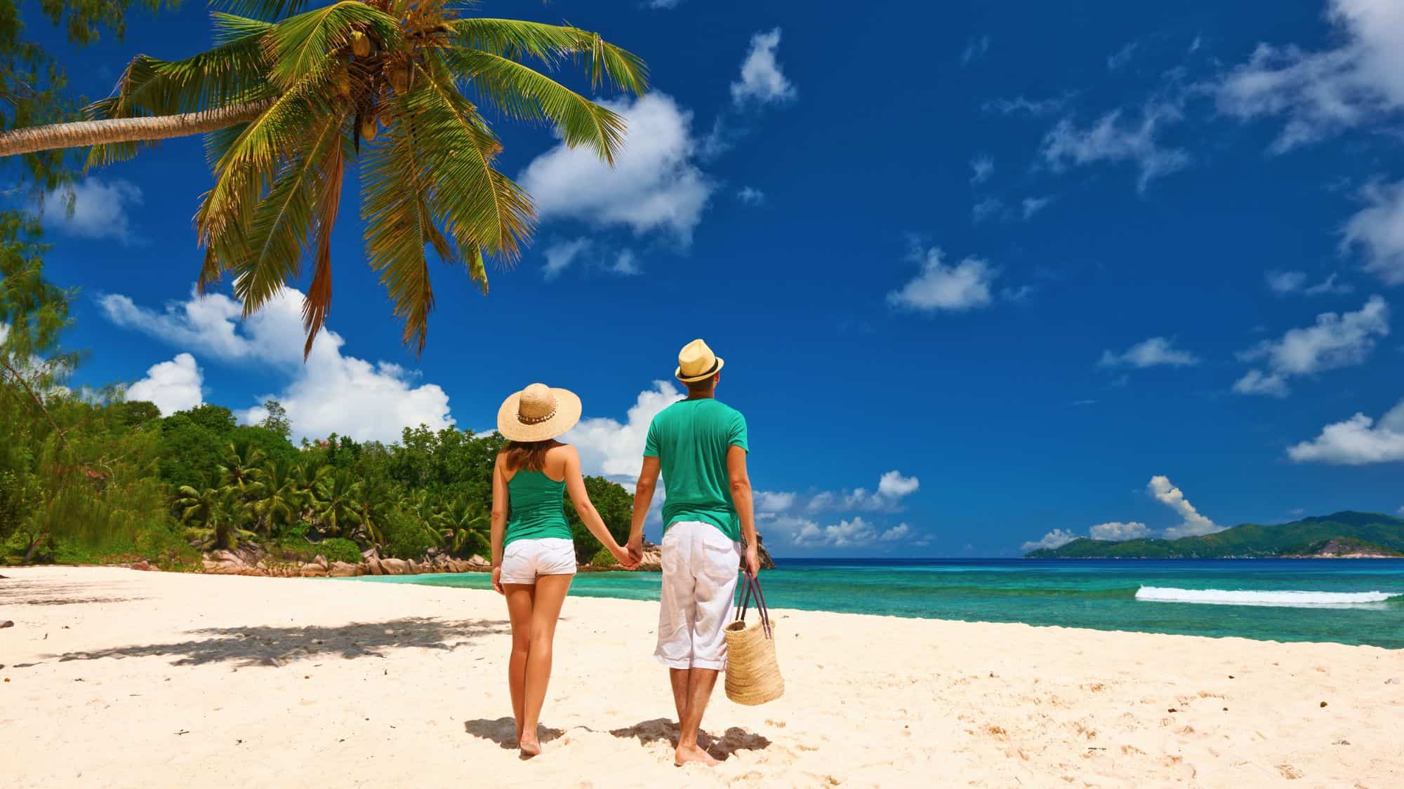 A couple strolls along a powdery white sand beach beneath leaning palms, with turquoise waters and lush jungle in the background.