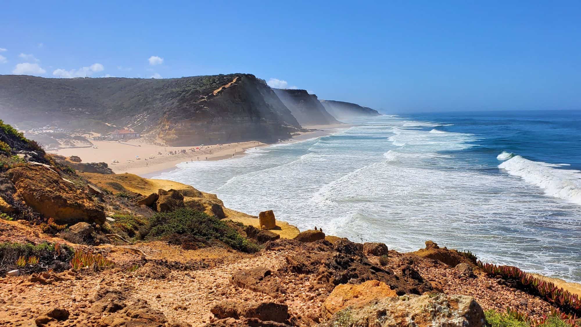 Golden cliffs drop steeply to a wide, sandy beach where waves crash ashore, with mist rising in the distance under a brilliant blue sky.